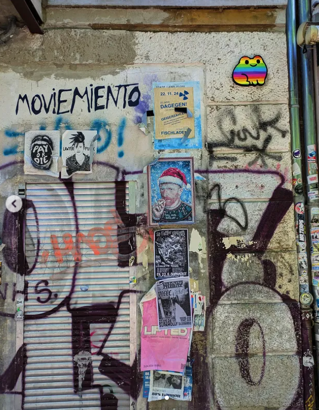 A graffiti-covered wall with various posters, stickers, and spray paint. The graffiti includes the words "MOVIMIENTO" and other tags, with some illustrations and a rainbow-colored cartoon sticker of a cute animal or character.