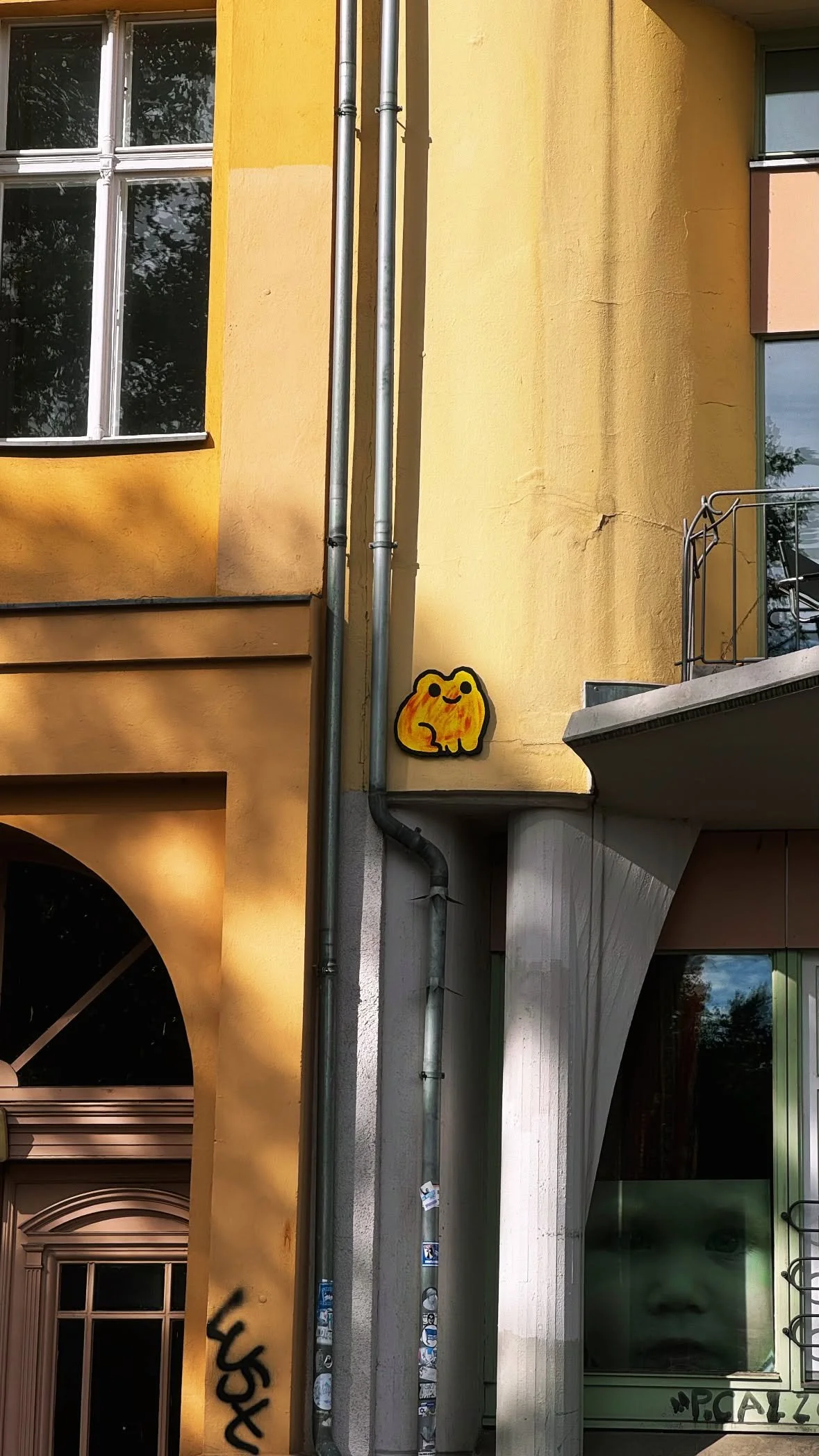 A yellow building with a corner ledge, several windows, and a small painted yellow, orange, and black cartoon-like character on the wall. There are also vertical metal pipes running up the side of the building and graffiti at the lower part.