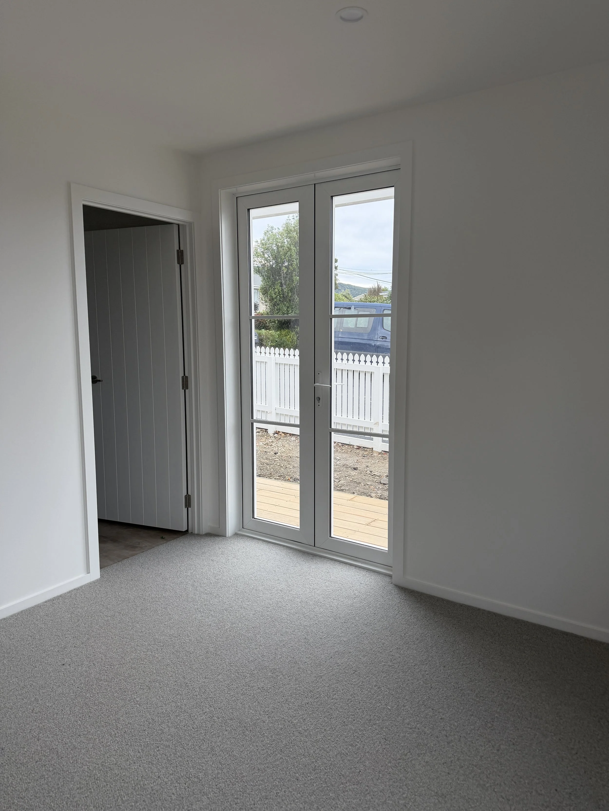 Empty room with gray carpet, white walls, a door on the left, and glass door leading outside to a small wooden deck and yard with a white fence and trees in the background.