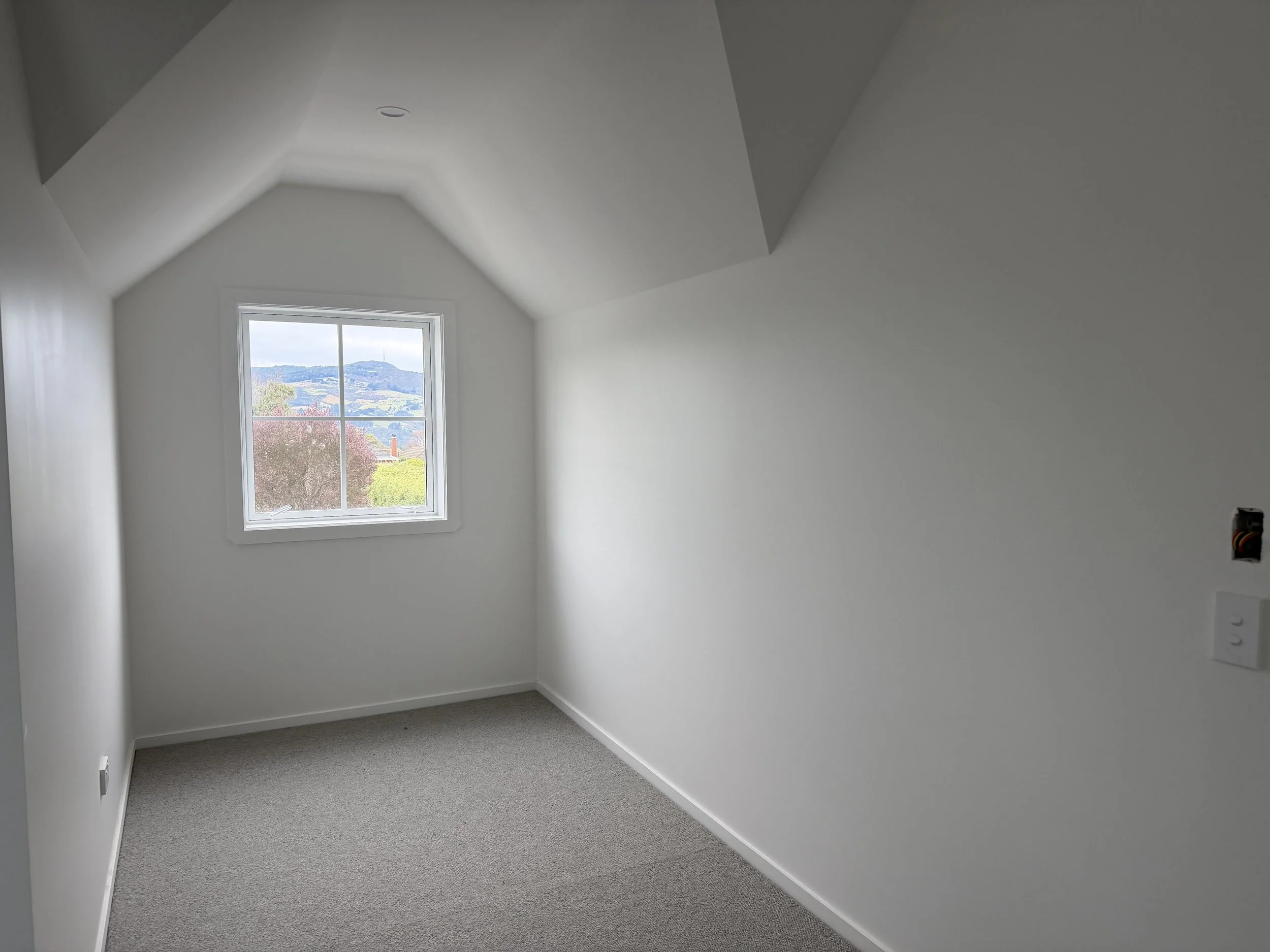 Empty white room with slanted ceiling, a window showing outdoor hills and trees, and a gray carpeted floor.