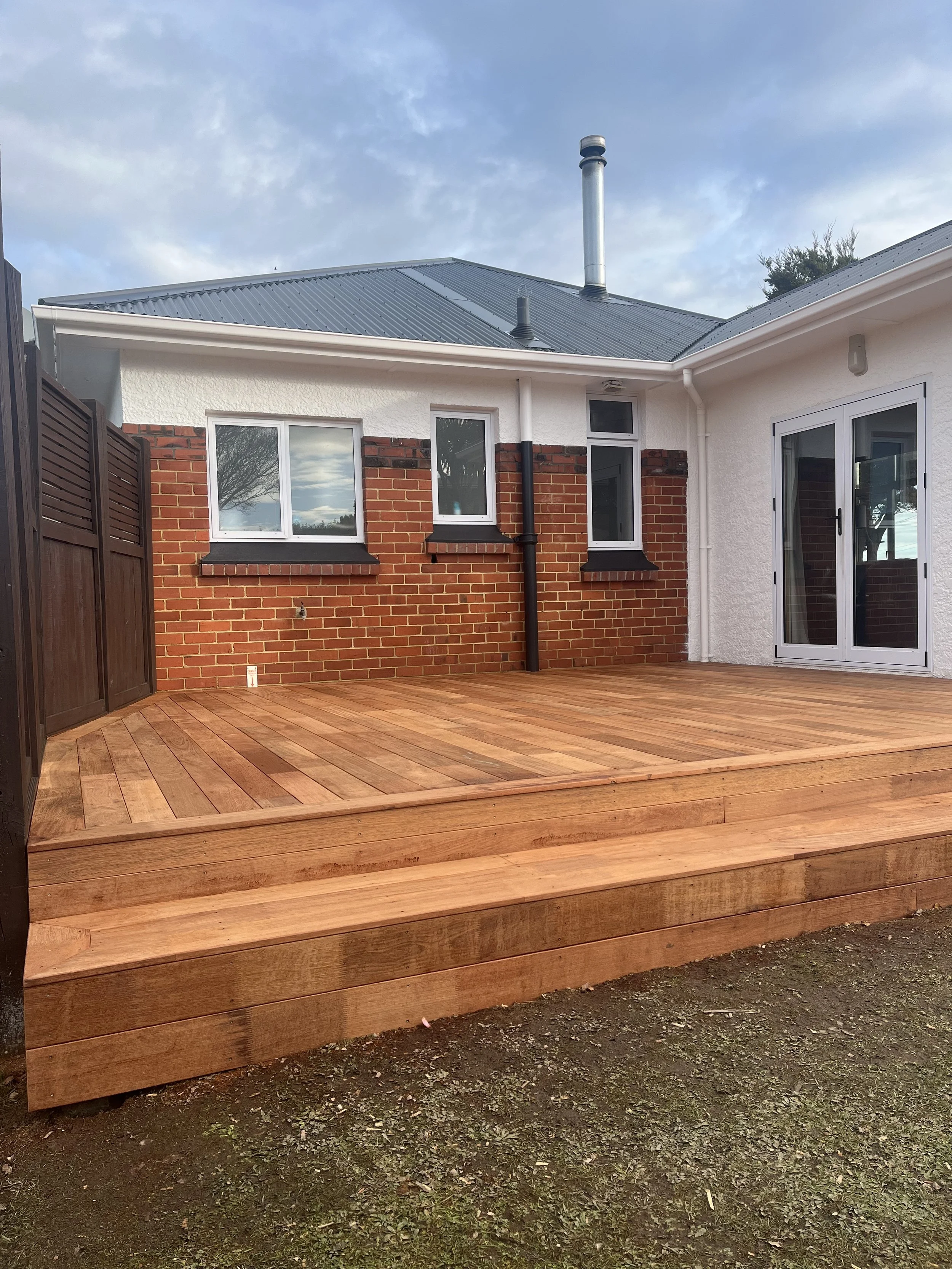 Photo of a backyard deck made of wooden planks with stairs, attached to a house with brick and white walls and sliding glass doors, on a cloudy day.