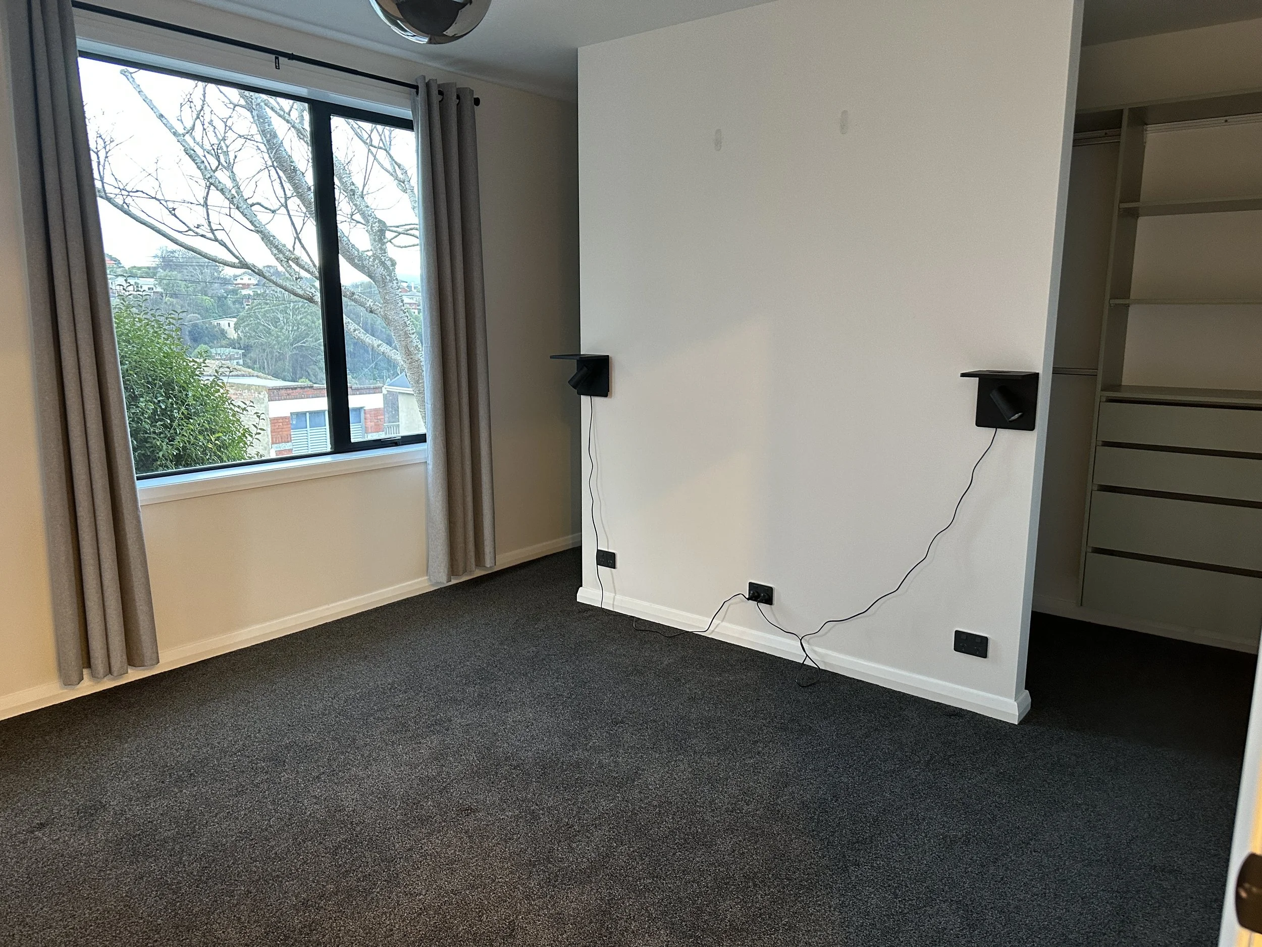 A corner of a room with a window showing trees and a neighborhood outside. There are two black speakers mounted on the white wall, with cables hanging down. The room has dark carpet flooring and white walls, with an open closet on the right side.