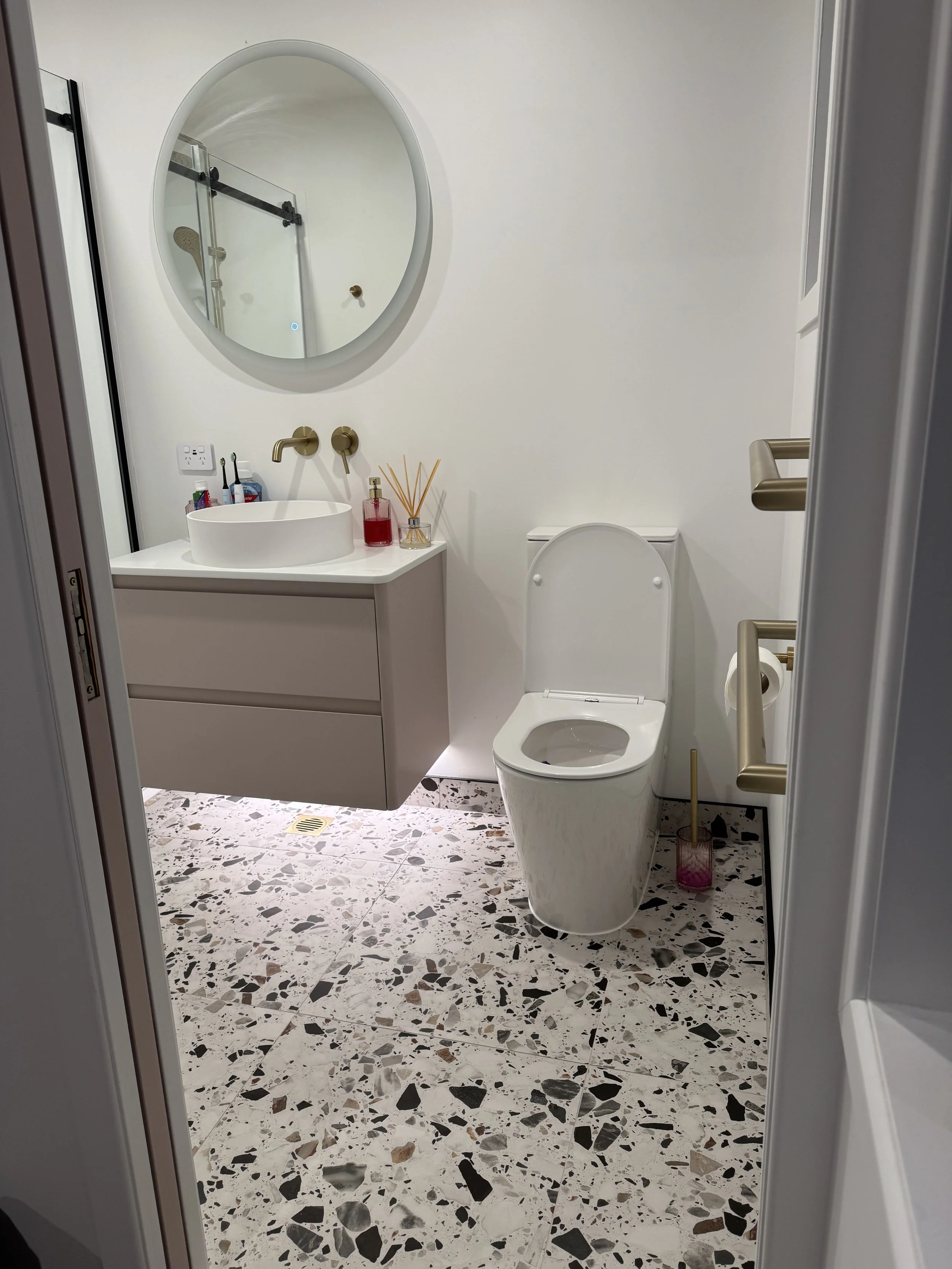 A modern bathroom with terrazzo flooring, white walls, a floating vanity with a basin sink, a gold faucet, toothbrushes, a soap dispenser, and reed diffusers. There's a wall-mounted toilet with a concealed cistern, a gold towel bar, and a large round