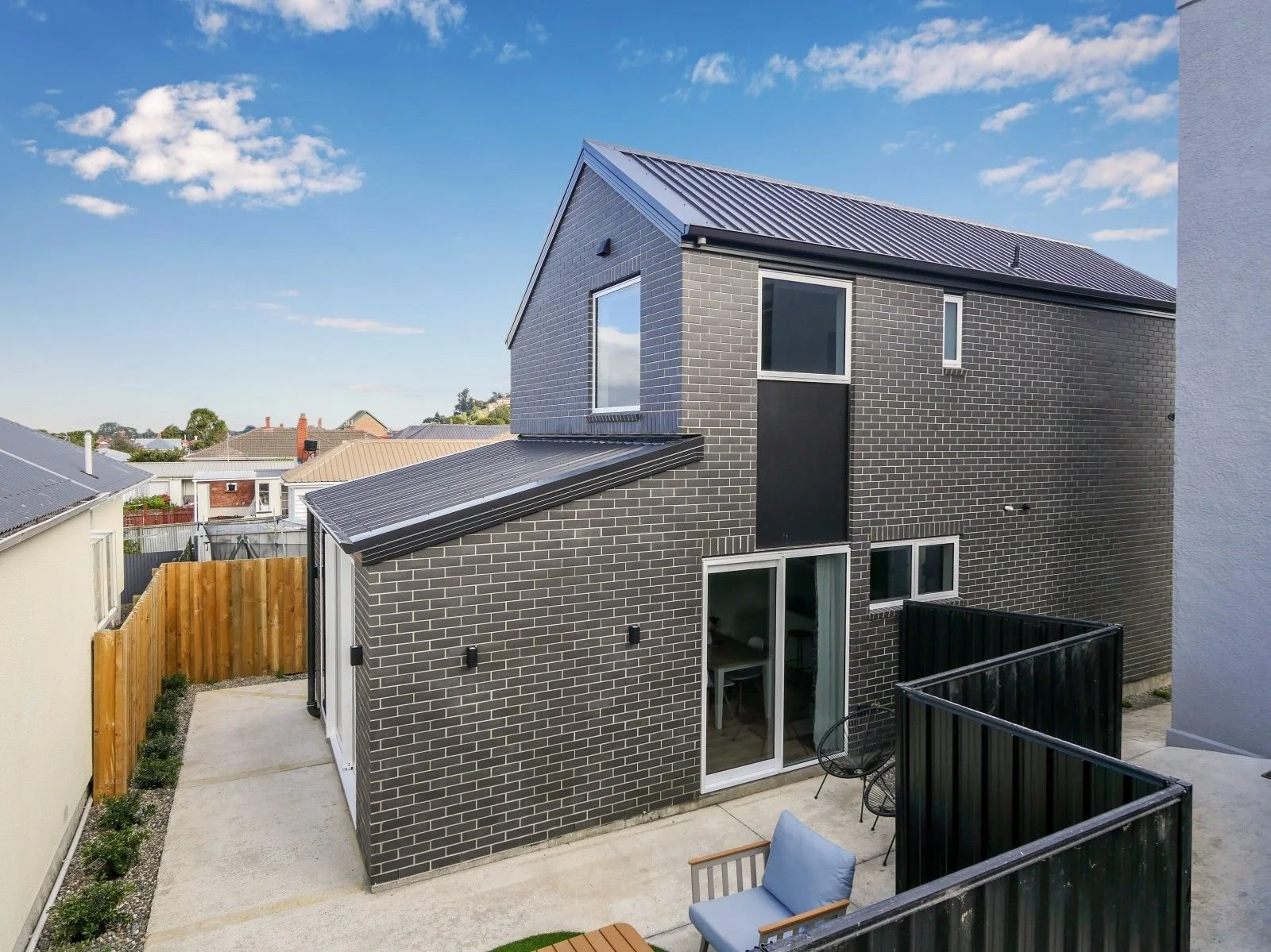 Modern two-story house with black brick exterior, metal roof, and a small backyard with patio furniture