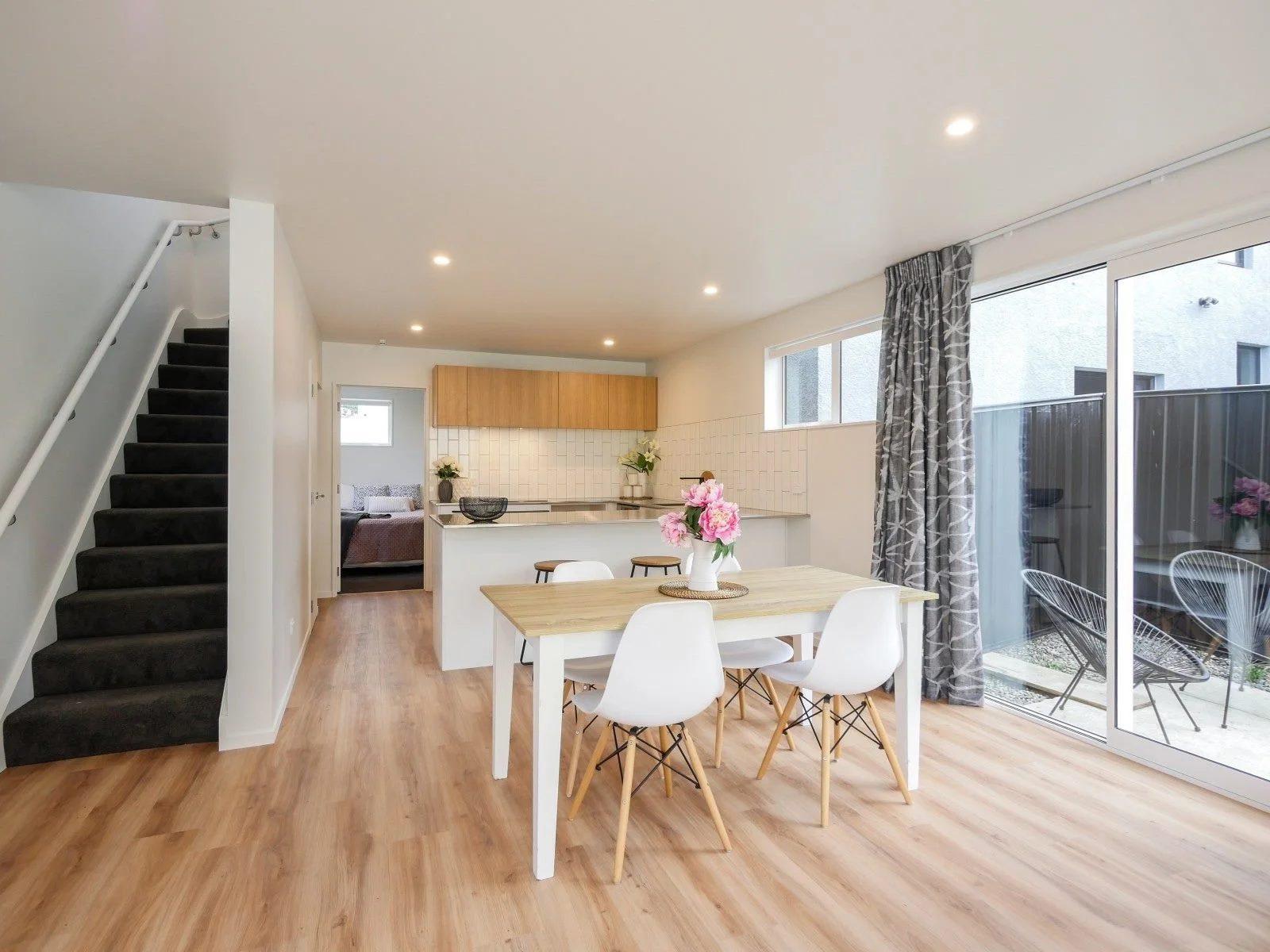 Open living and dining area with wooden flooring, white walls, a glass sliding door to an outdoor balcony, a dining table with white chairs, a kitchen with wooden cabinets, a breakfast bar, and decorative flowers.