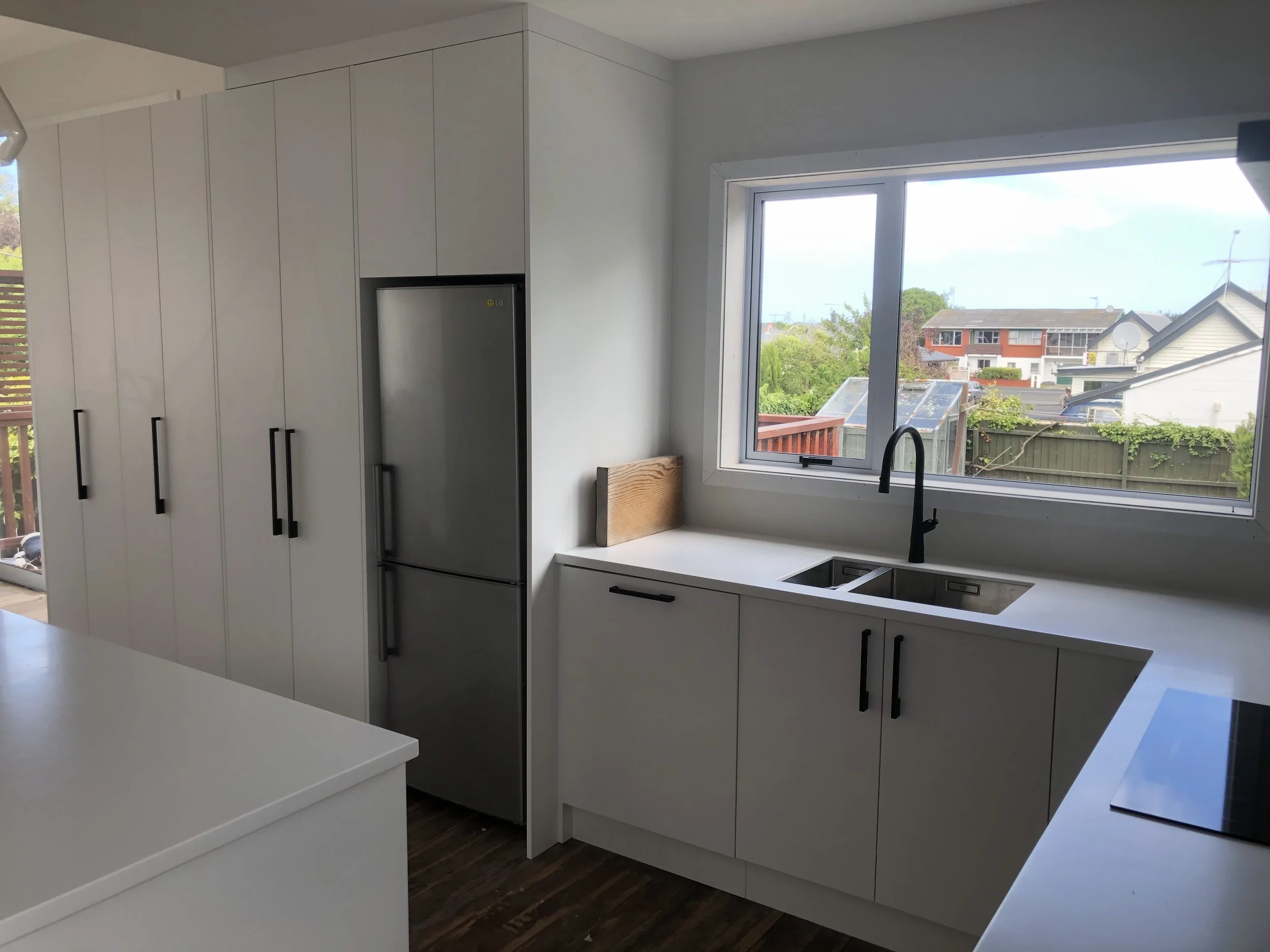 A modern kitchen with white cabinets, a large window overlooking a neighborhood, and a stainless steel refrigerator.