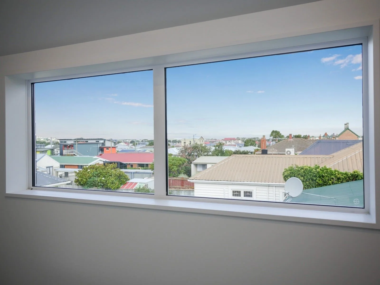 View through a large window showing a neighborhood with houses, rooftops, trees, and a partly cloudy blue sky.