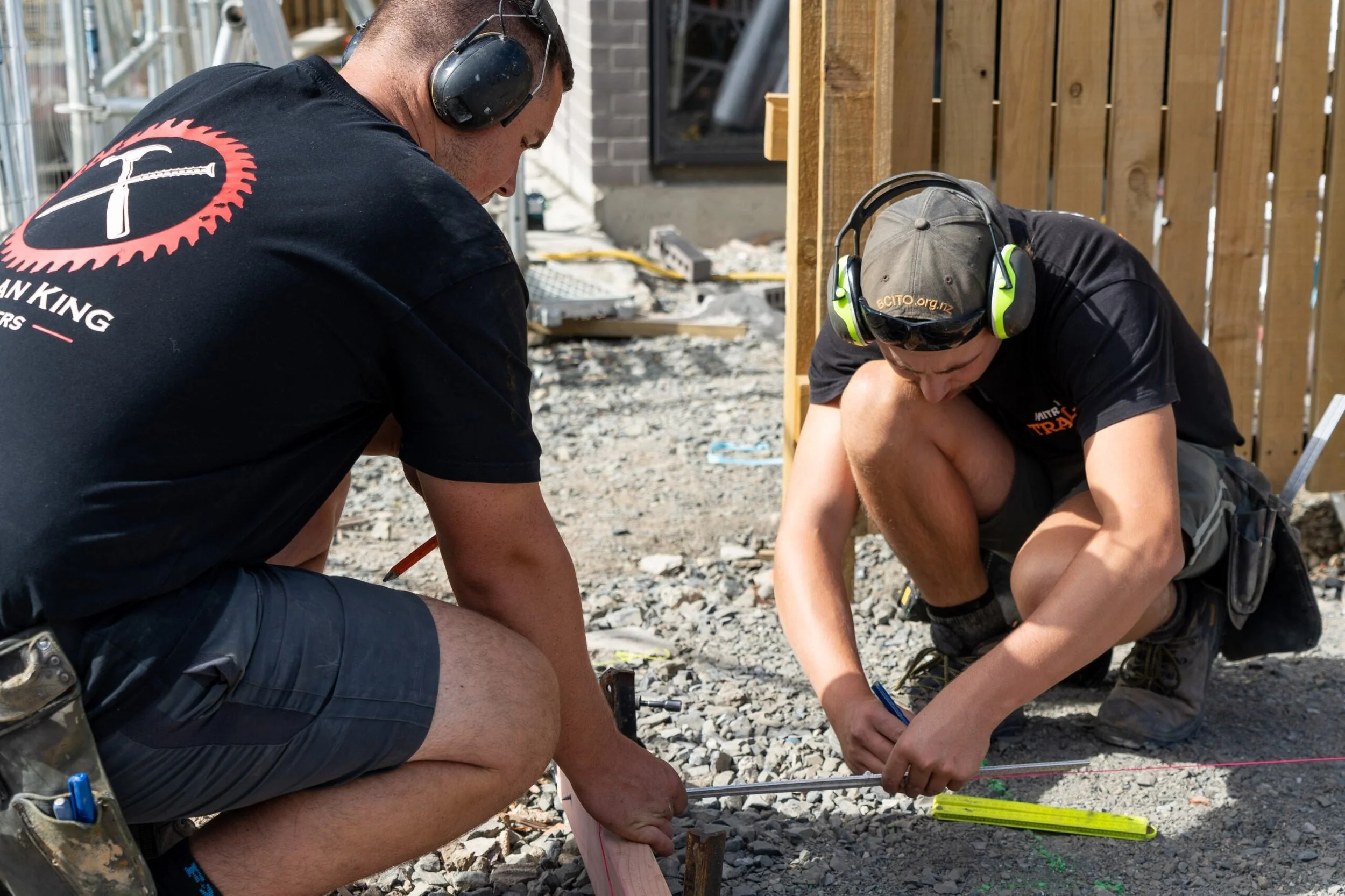 Two construction workers, wearing protective headphones, are working on a construction site with wooden fencing in the background. They are kneeling on gravel, using measuring tools on the ground.