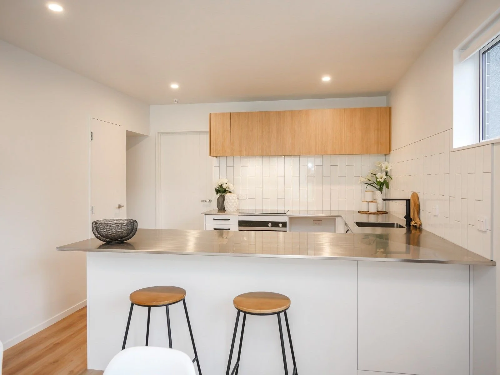 Modern kitchen with white walls, wooden cabinets, black sink, white tiled backsplash, and a stainless steel countertop island with two wooden bar stools.