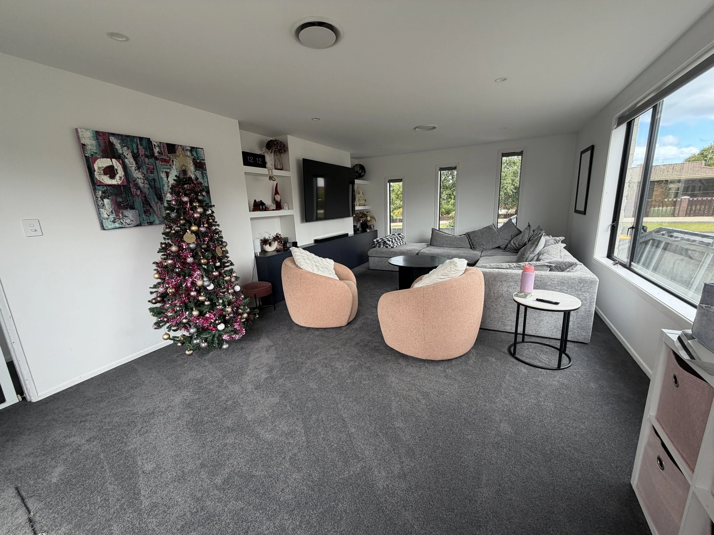Living room with gray sectional sofa, pink armchairs, Christmas tree, wall art, chalkboard shelves, windows with view of outdoors, and various decorative items.