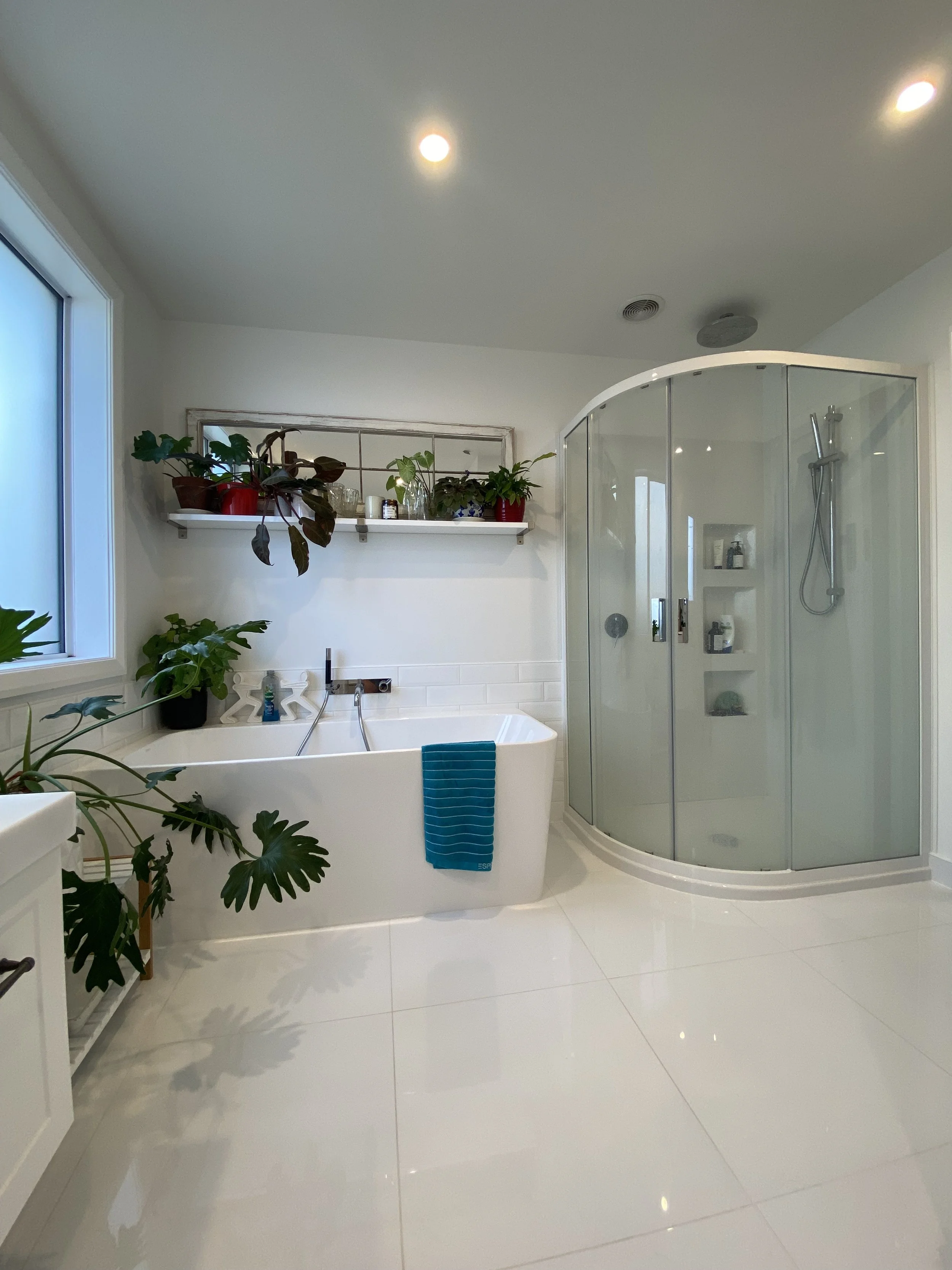 Modern bathroom with white tiles, a bathtub with a turquoise towel, glass shower enclosure, and potted plants on a shelf and floor.