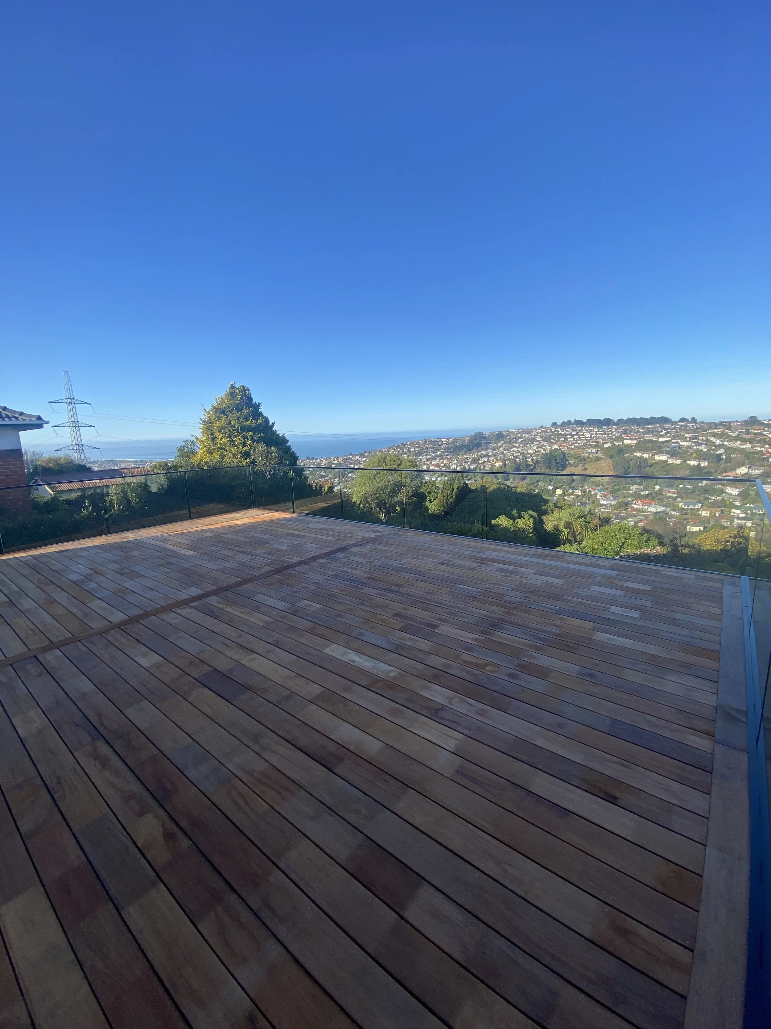 A spacious outdoor wooden deck with glass railing, overlooking a hilly suburban area with houses, trees, and a distant sea under a clear blue sky.
