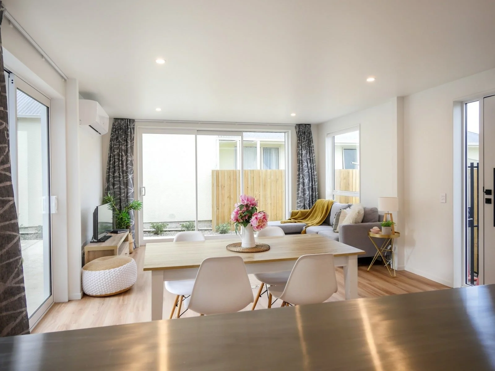 Bright and airy living room with a dining area, large sliding glass doors, a small TV on a wooden stand, decorative plants, a gray couch with yellow and white pillows, a table lamp, and colorful flowers in a vase.