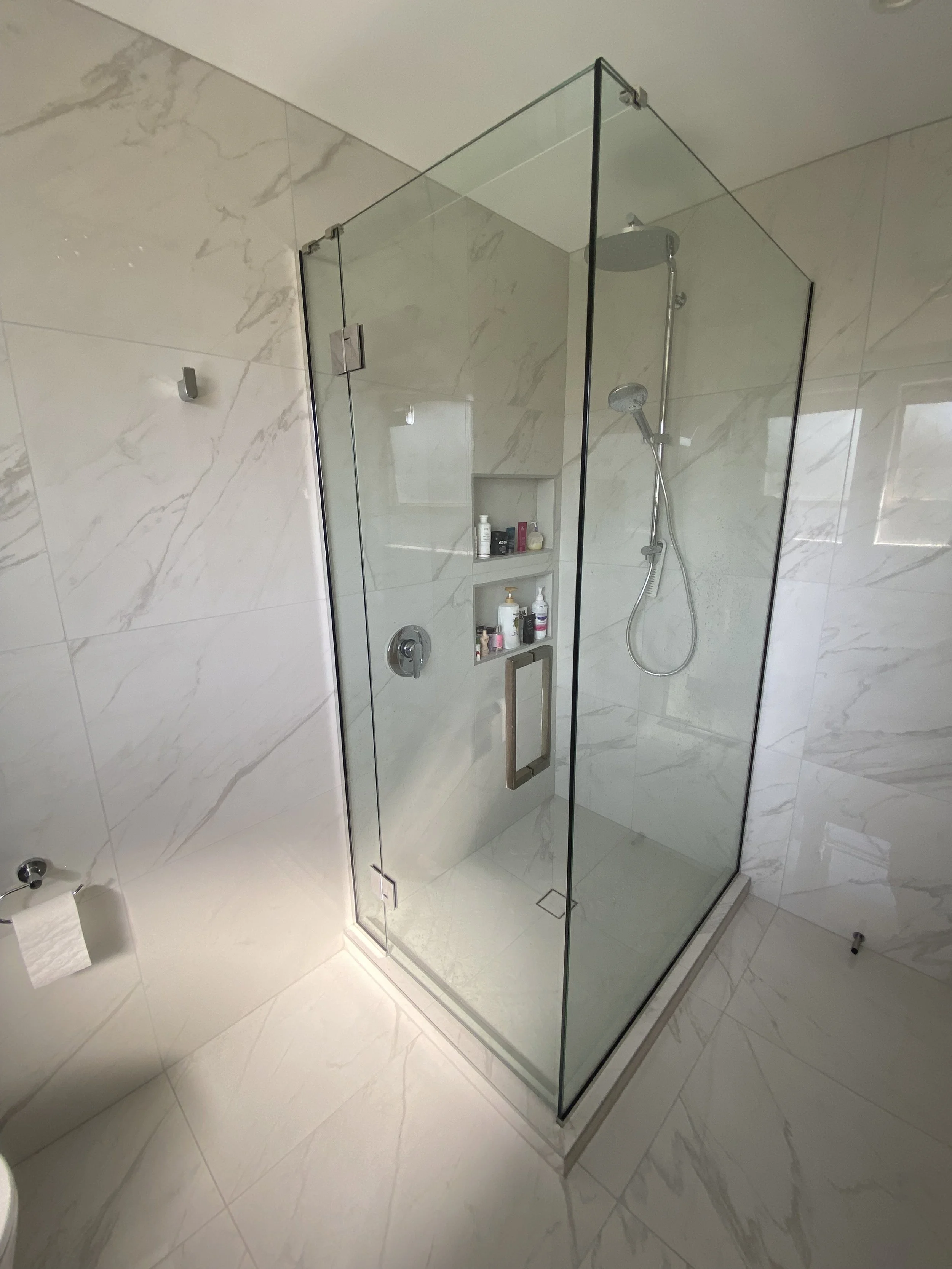A modern shower with a glass enclosure, marble tiles, a wall niche with shampoo and soap bottles, and a handheld showerhead.
