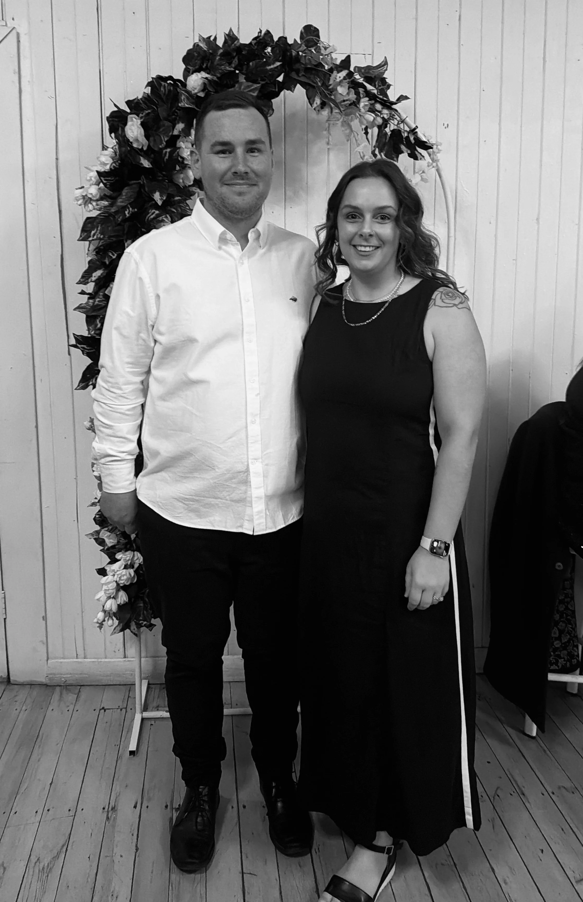 A man and woman standing together indoors, smiling for the camera, with a floral wreath in the background.