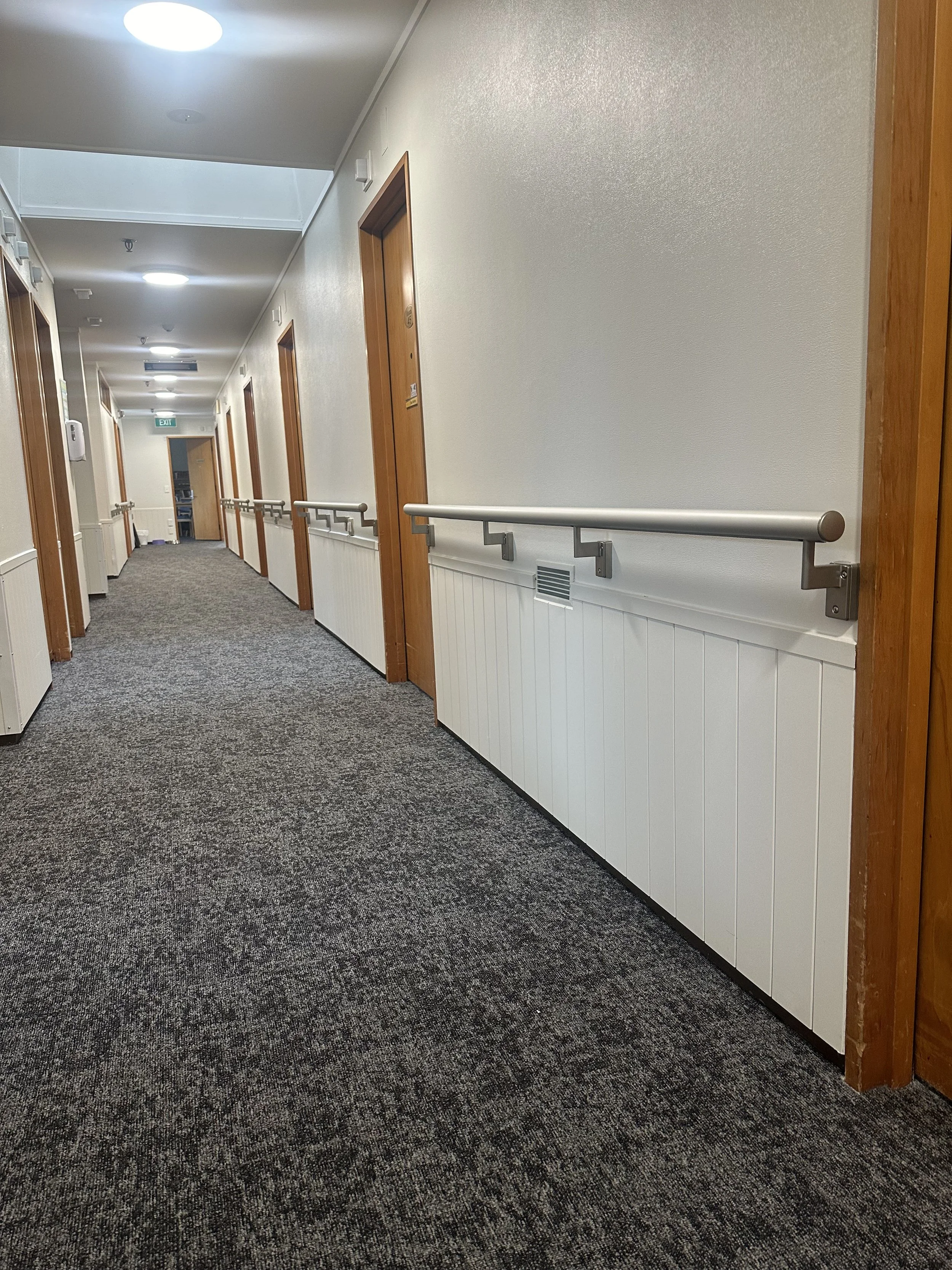 An indoor corridor with carpeted flooring, wooden door frames, a handrail along the wall, and ceiling lights.