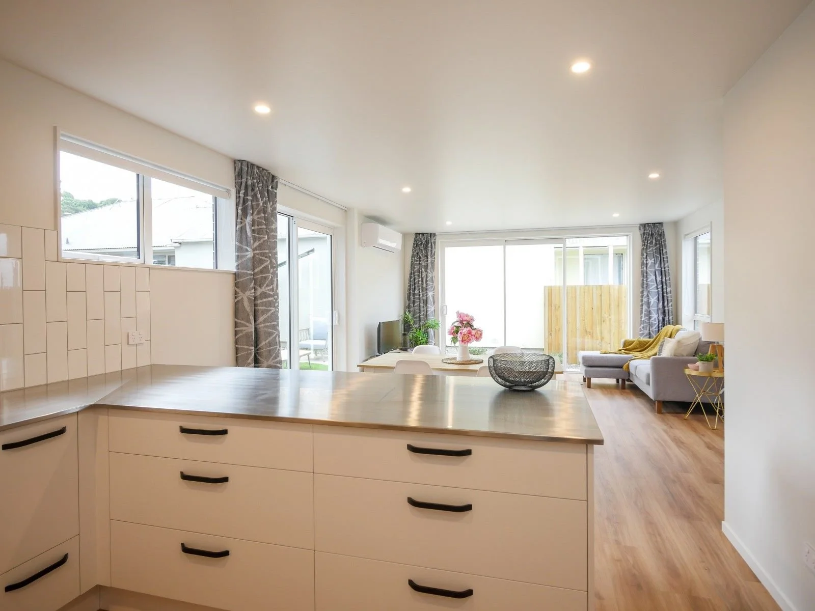 Open-concept living room and kitchen with large windows, a gray sofa, a small TV, and a dining table with flowers.