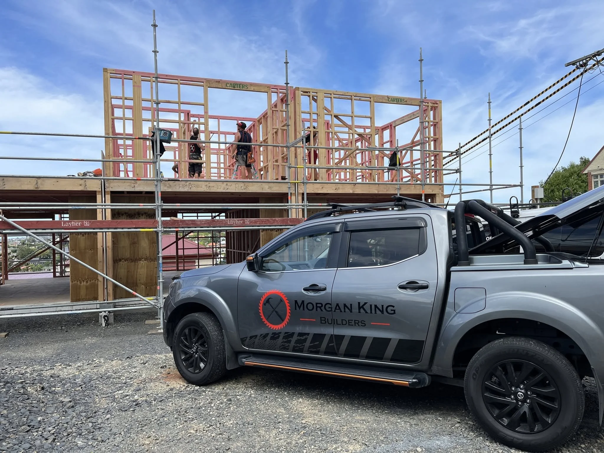 Construction workers working on the wooden frame of a building under a blue sky, with a gray vehicle parked in front and scaffolding surrounding the site.