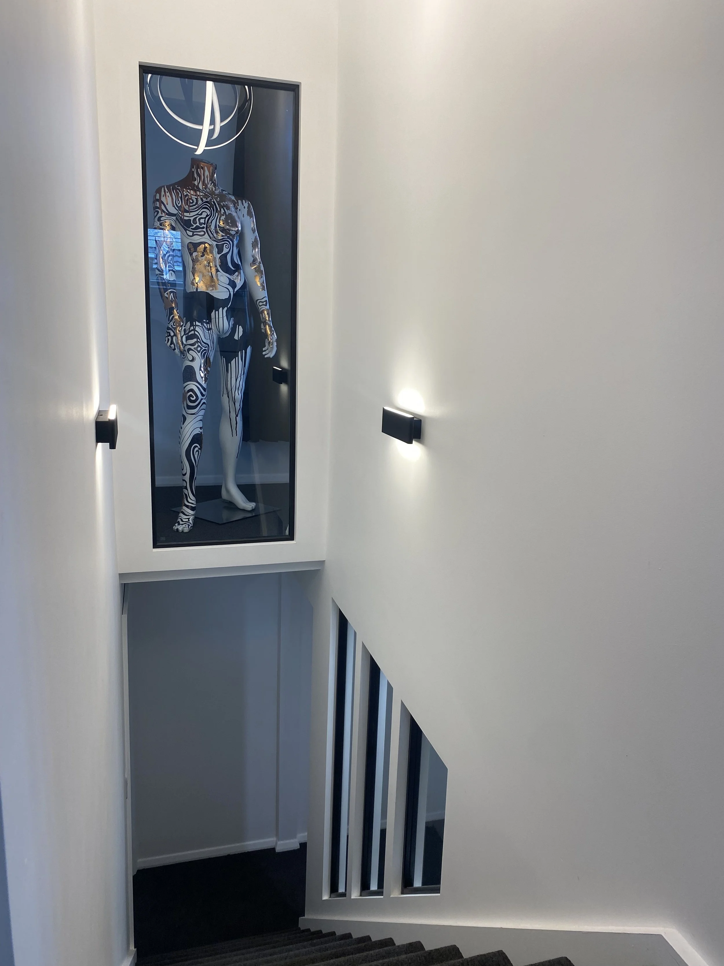 A mannequin inside a display case with black, white, and gold patterns, located at the top of a modern staircase in a bright interior with white walls and black carpeted stairs.