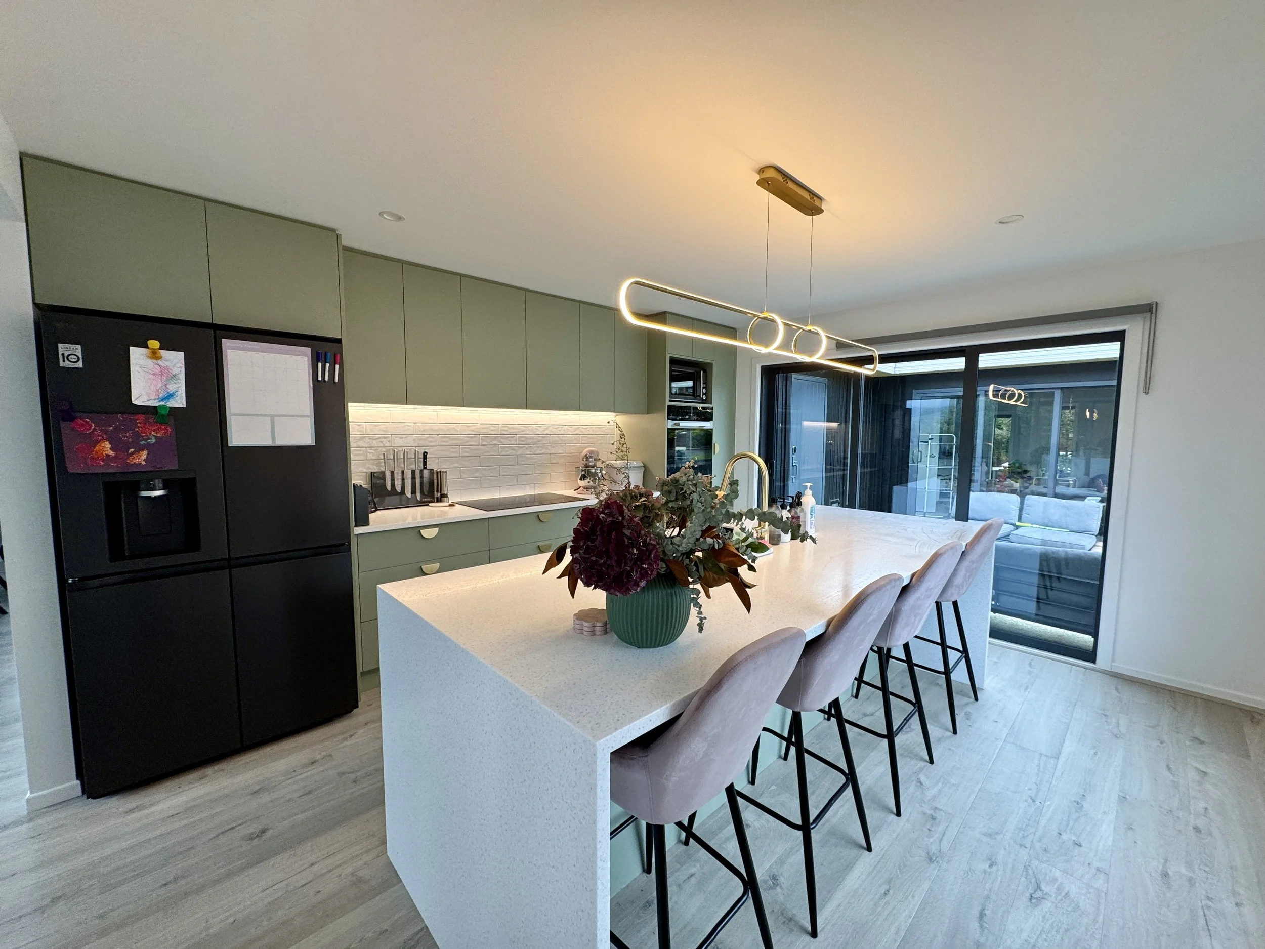 Modern kitchen with green cabinets, white countertop, black refrigerator, flowers on the island, and sitting area with large windows.