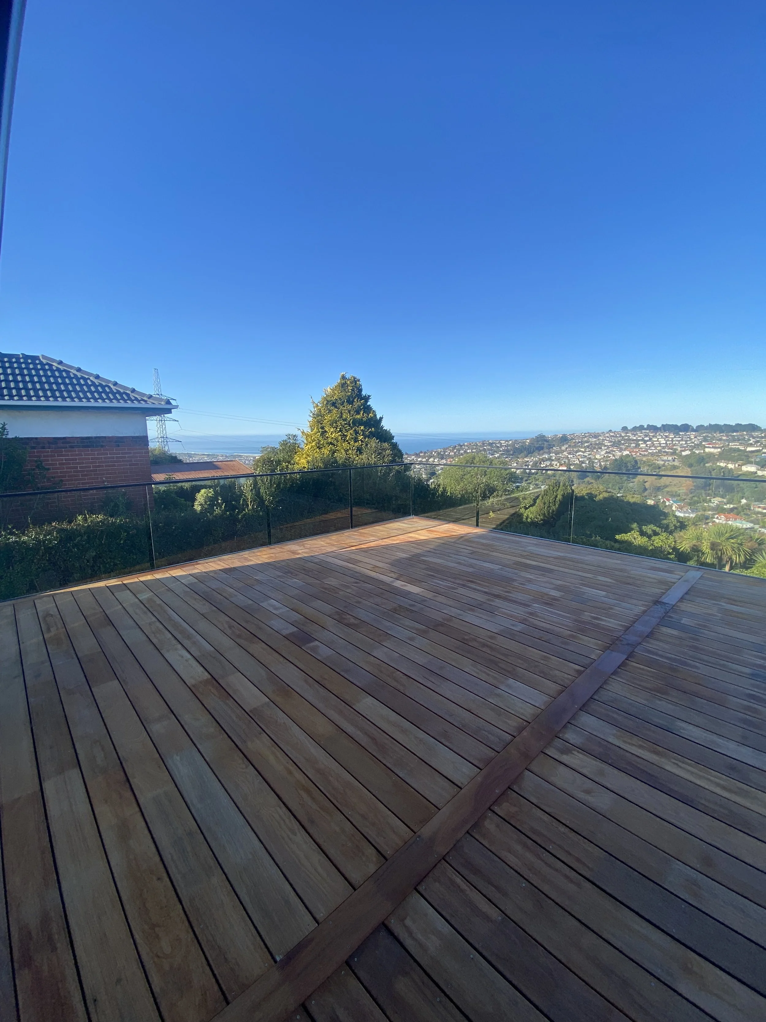 View from a wooden deck with glass railing, overlooking a suburban neighborhood, trees, and distant hills under a clear blue sky.