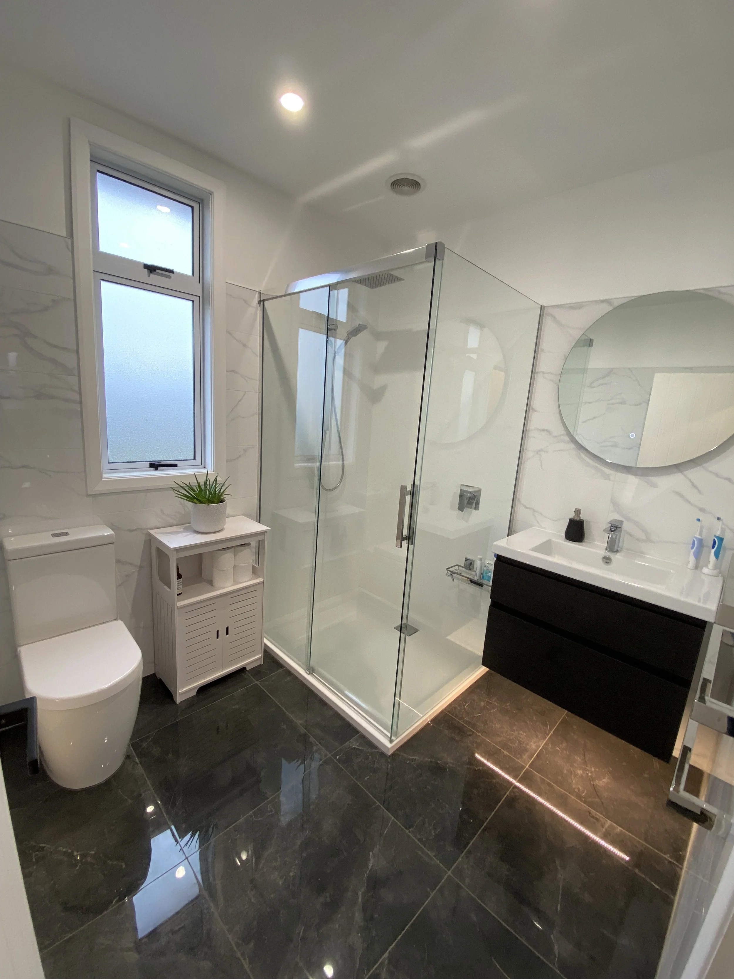 Modern bathroom with black marble floor, white marble walls, a glass shower enclosure, a white toilet, a small white storage unit with a potted plant, a sink with a black cabinet, round mirrors, and a window letting in natural light.