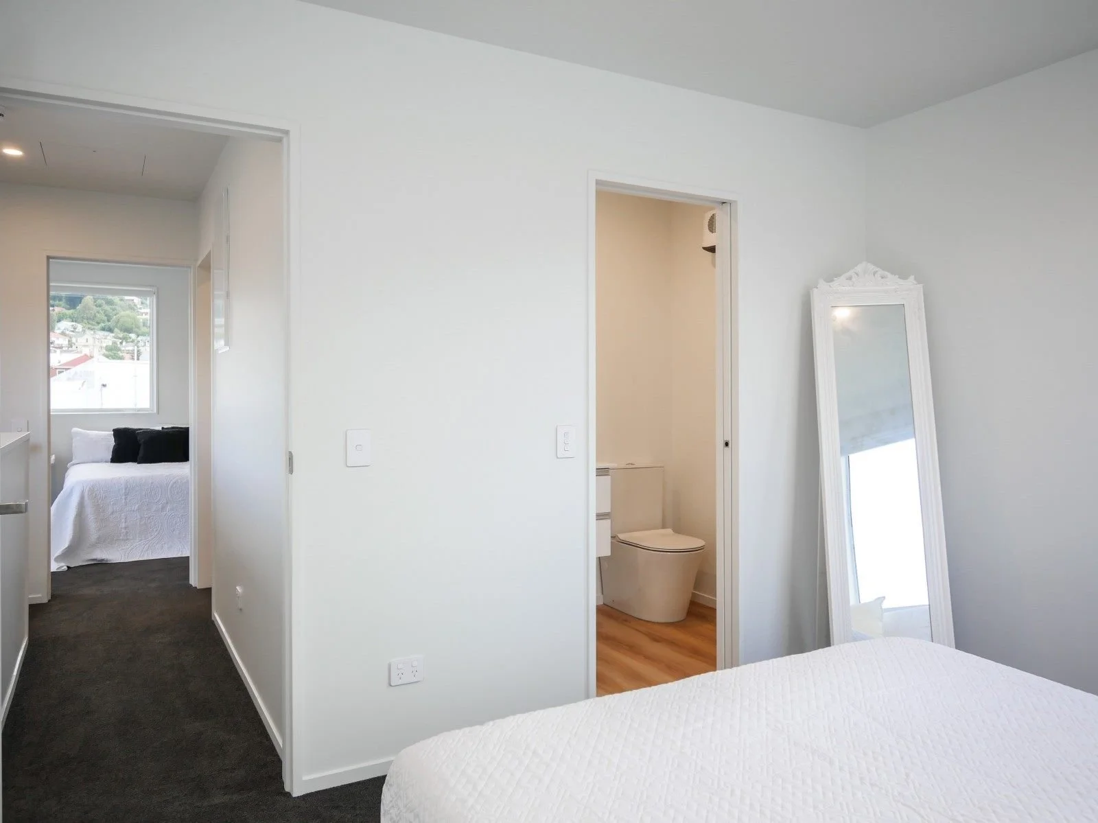 Interior view of a modern apartment with white walls, a mirror leaning against the wall, a visible bathroom with a toilet, and a bedroom with a bed and window.