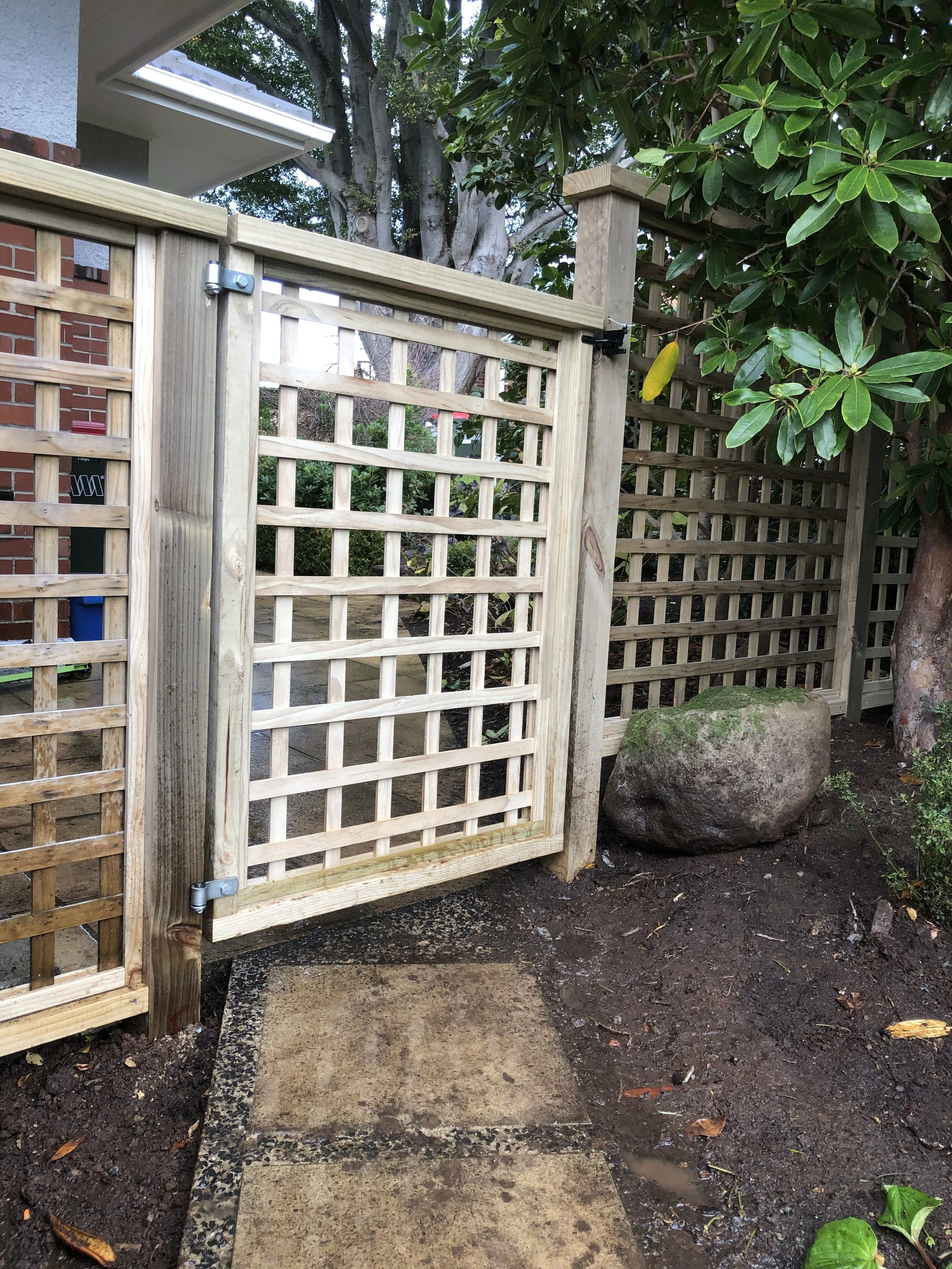 A wooden gate with lattice design, attached to a fence, next to a rock and a small patch of dirt with some green plants, outside near trees and a house.