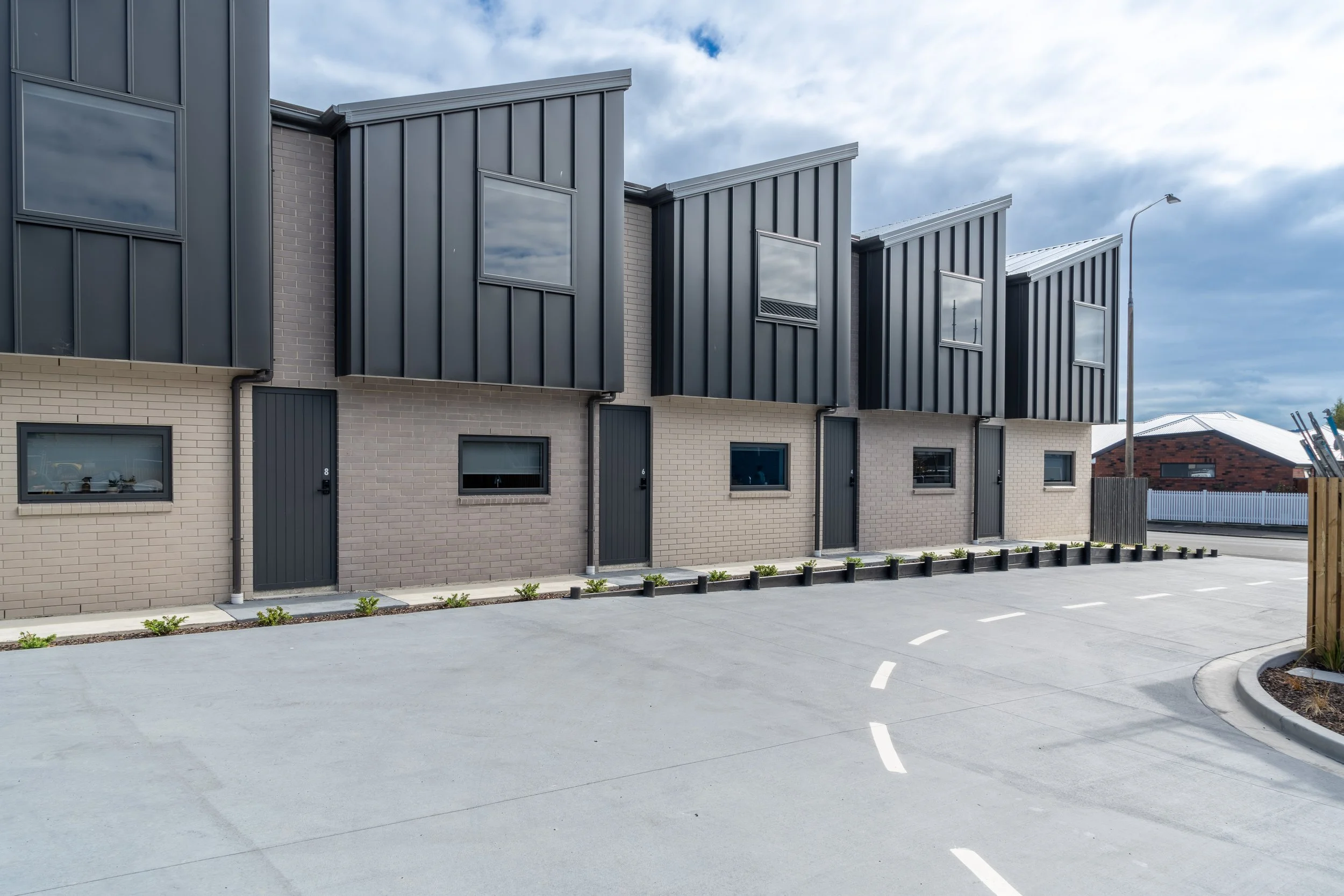 Exterior of modern row houses with black metal siding and brick lower levels, small windows, and a paved parking area with designated parking spots.