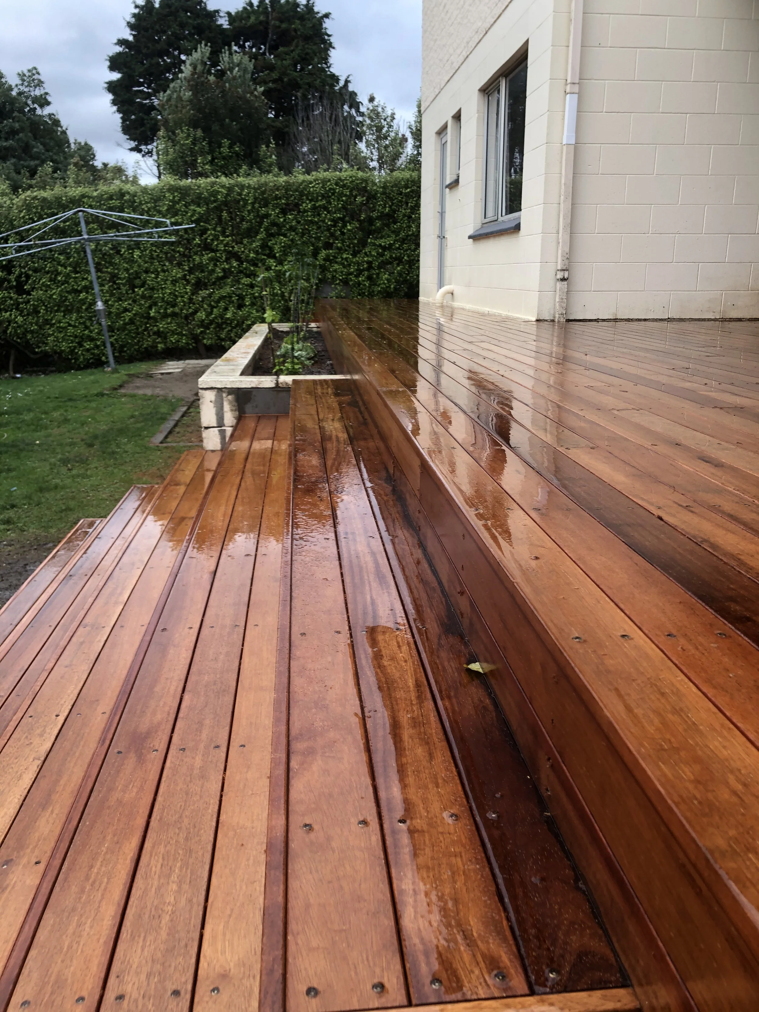 Wet wooden deck attached to a house, with a garden, hedge, and trees in the background on a cloudy day.