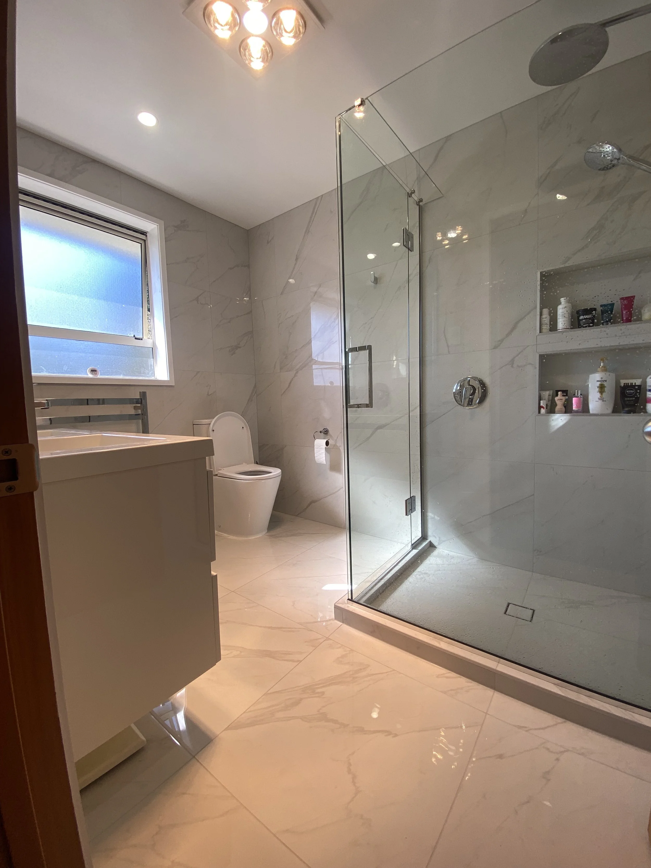 A modern bathroom with frosted window, large marble tiles, a glass shower enclosure with a showerhead, built-in shelves with toiletries, a floating vanity with a sink, and a toilet.
