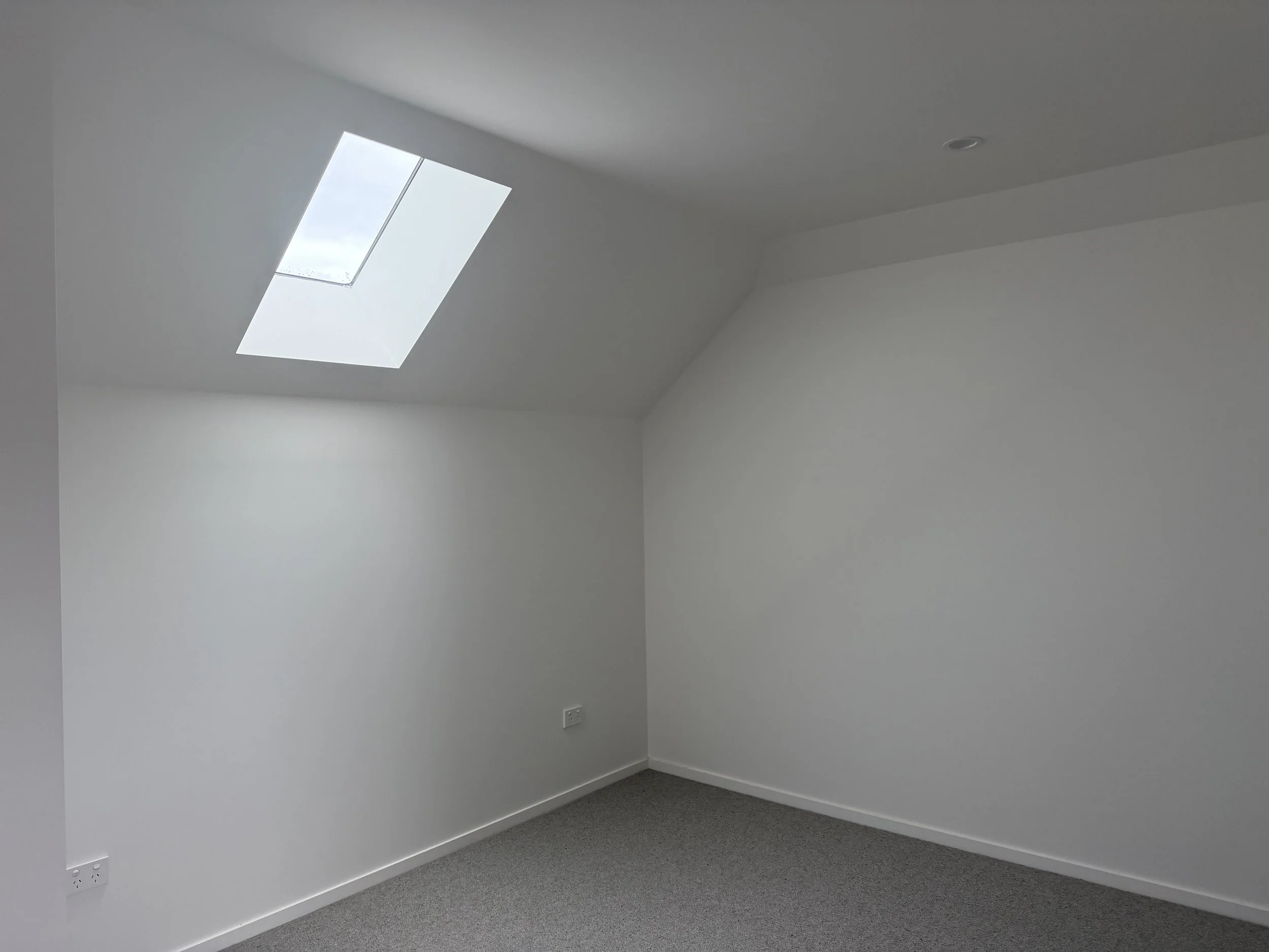 Empty white room with a sloped ceiling and skylight window, gray carpeted floor, electrical outlets, and a ceiling light