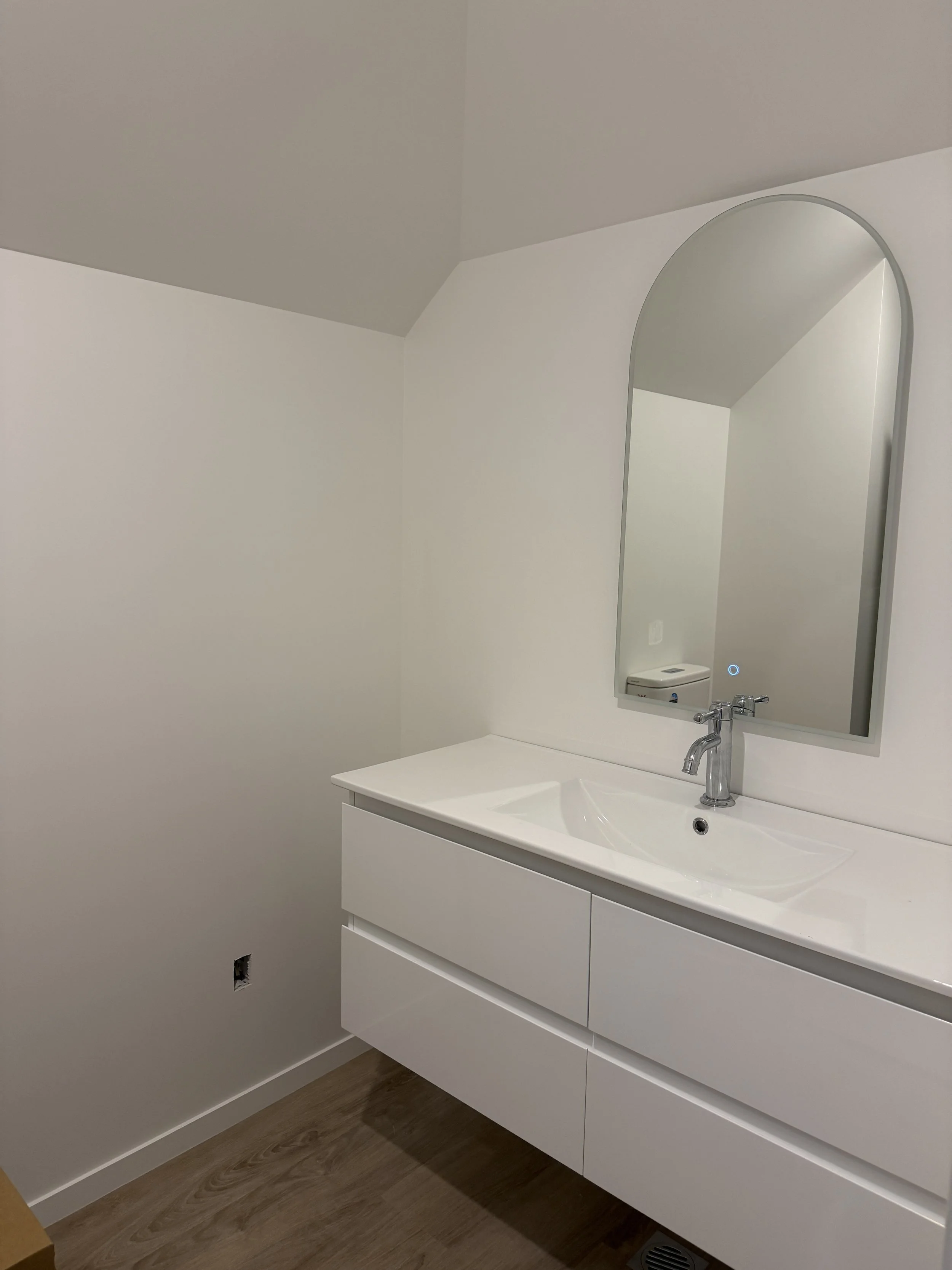Modern bathroom with white vanity, mirror, faucet, and wooden floor.