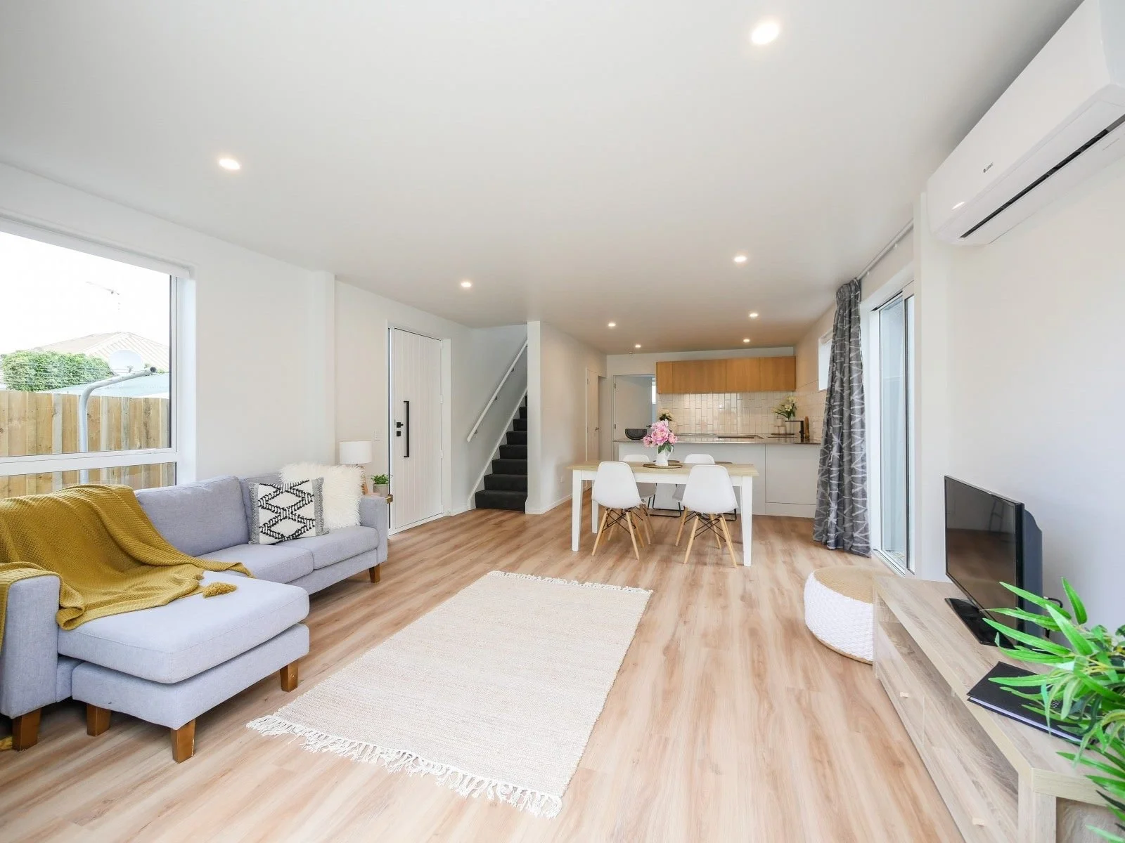 Open-concept living room and kitchen with light wood flooring, white walls, large windows, gray sofa with yellow blanket, white dining table, and modern kitchen in background.