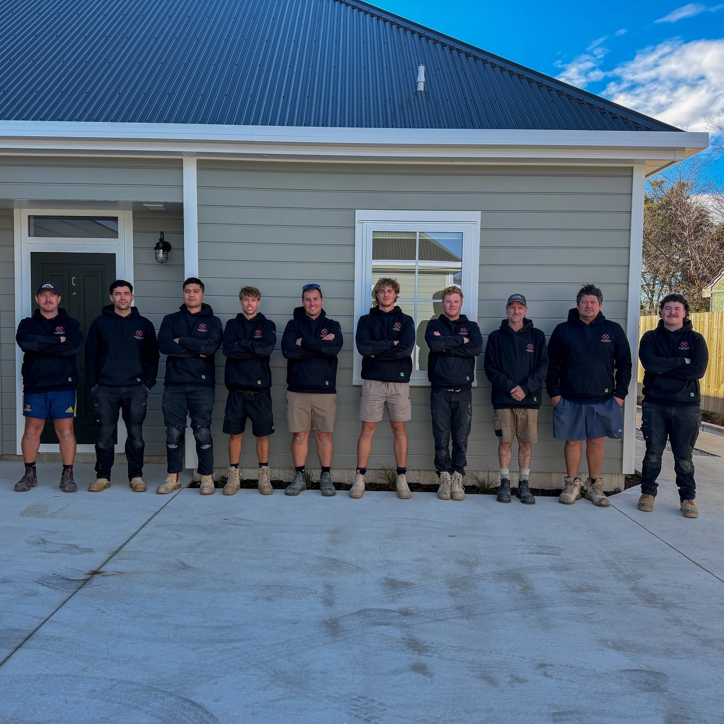 Group of people standing outside in front of a house with a metal roof, posing for a photo on a sunny day.