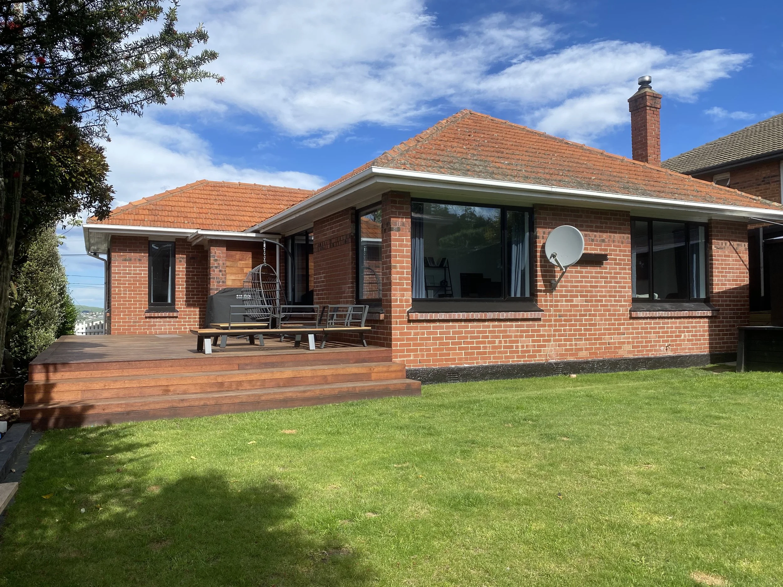 A brick house with large windows and a wooden deck in the backyard. There are outdoor chairs and a satellite dish attached to the house. The sky is partly cloudy.