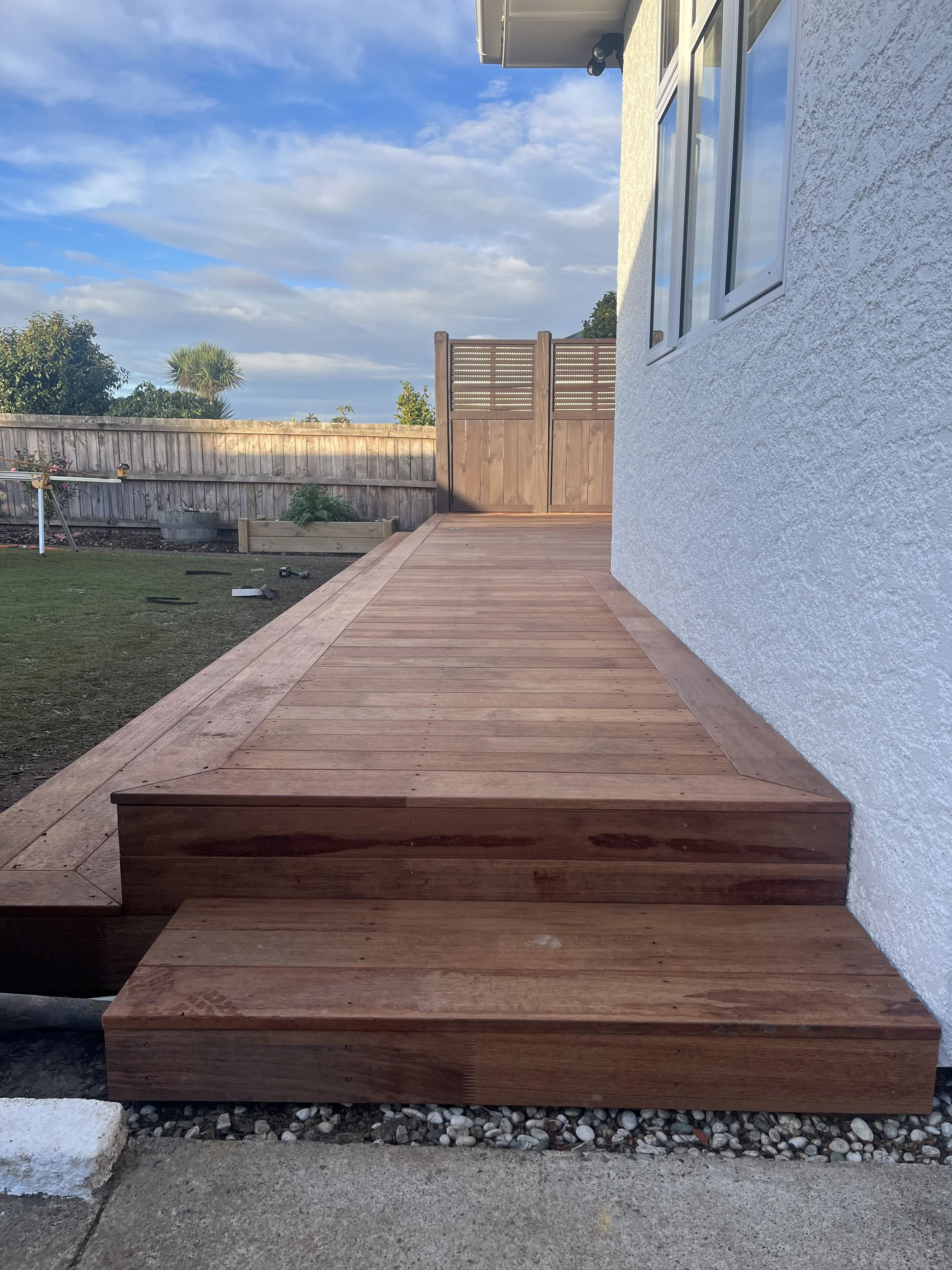Newly built wooden deck with stairs attached to the back of a house, white stucco exterior, a window, and a privacy fence in the background, under a partly cloudy sky.