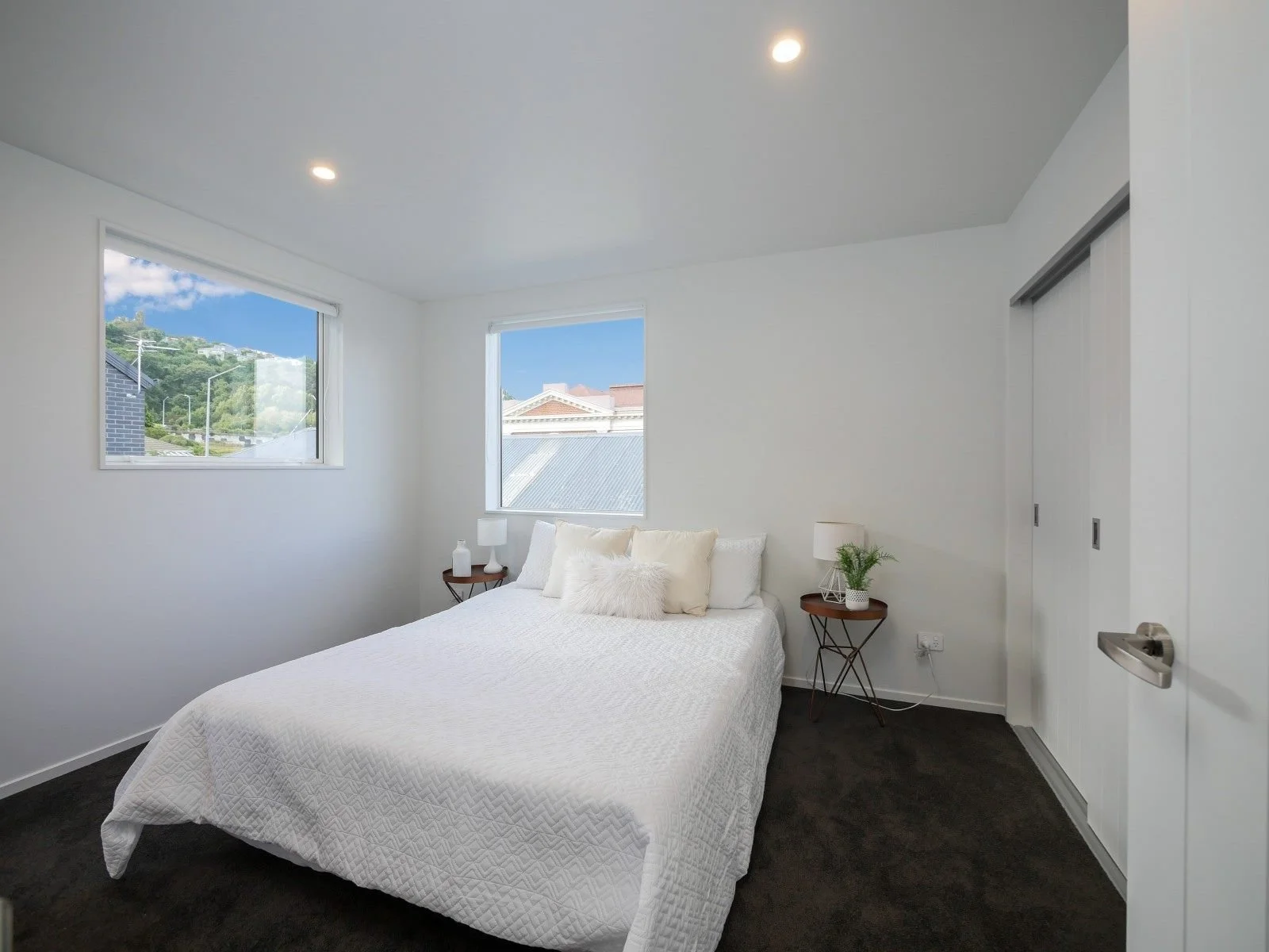 A bedroom with white walls, a bed with white bedding and several white pillows, two small round wooden nightstands with lamps and plants, two windows with a view of a neighborhood, and a sliding closet door.