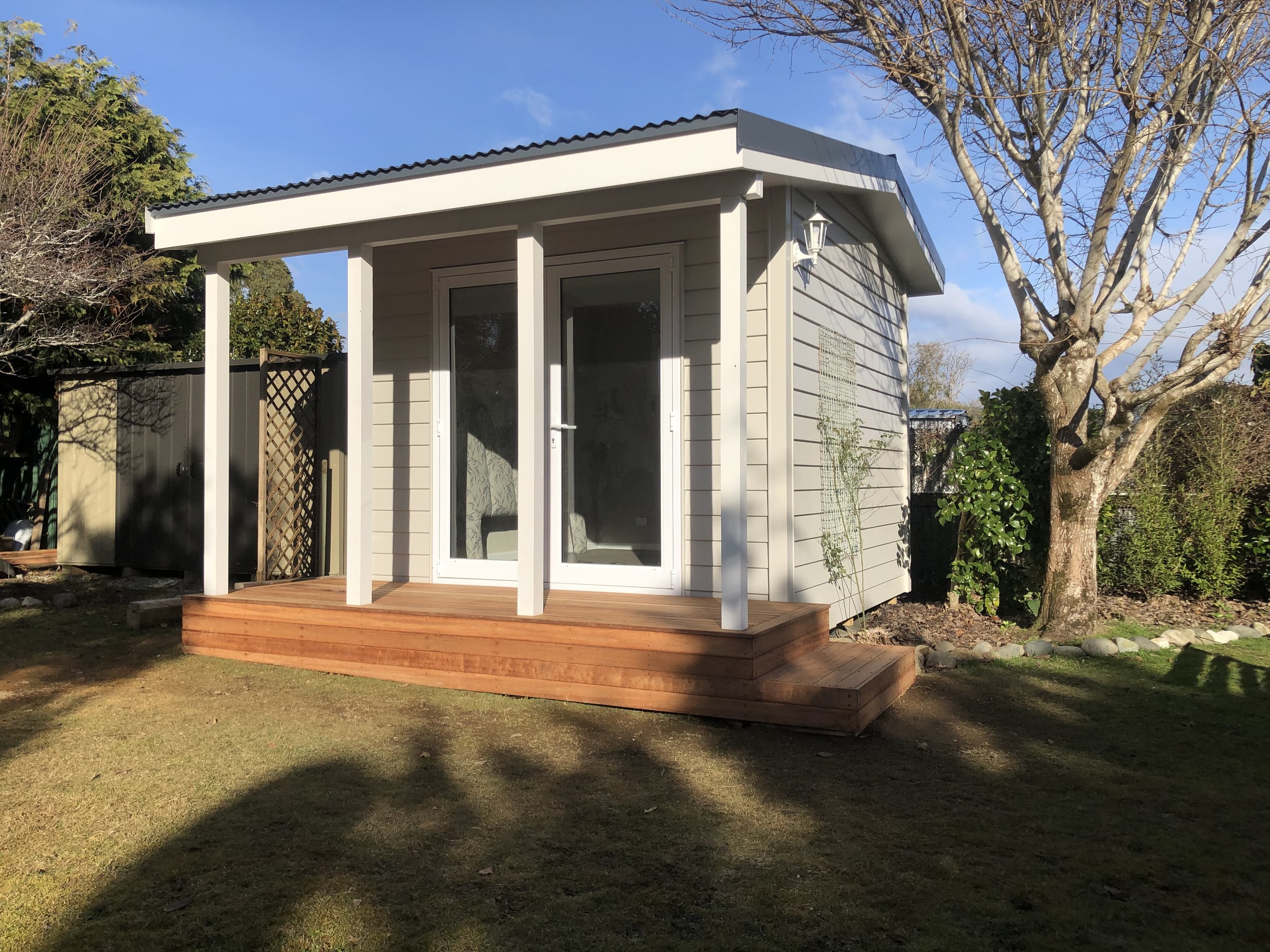 Small beige house with a wooden deck, sliding glass door, and outdoor lantern, surrounded by trees and a grassy yard on a clear day.