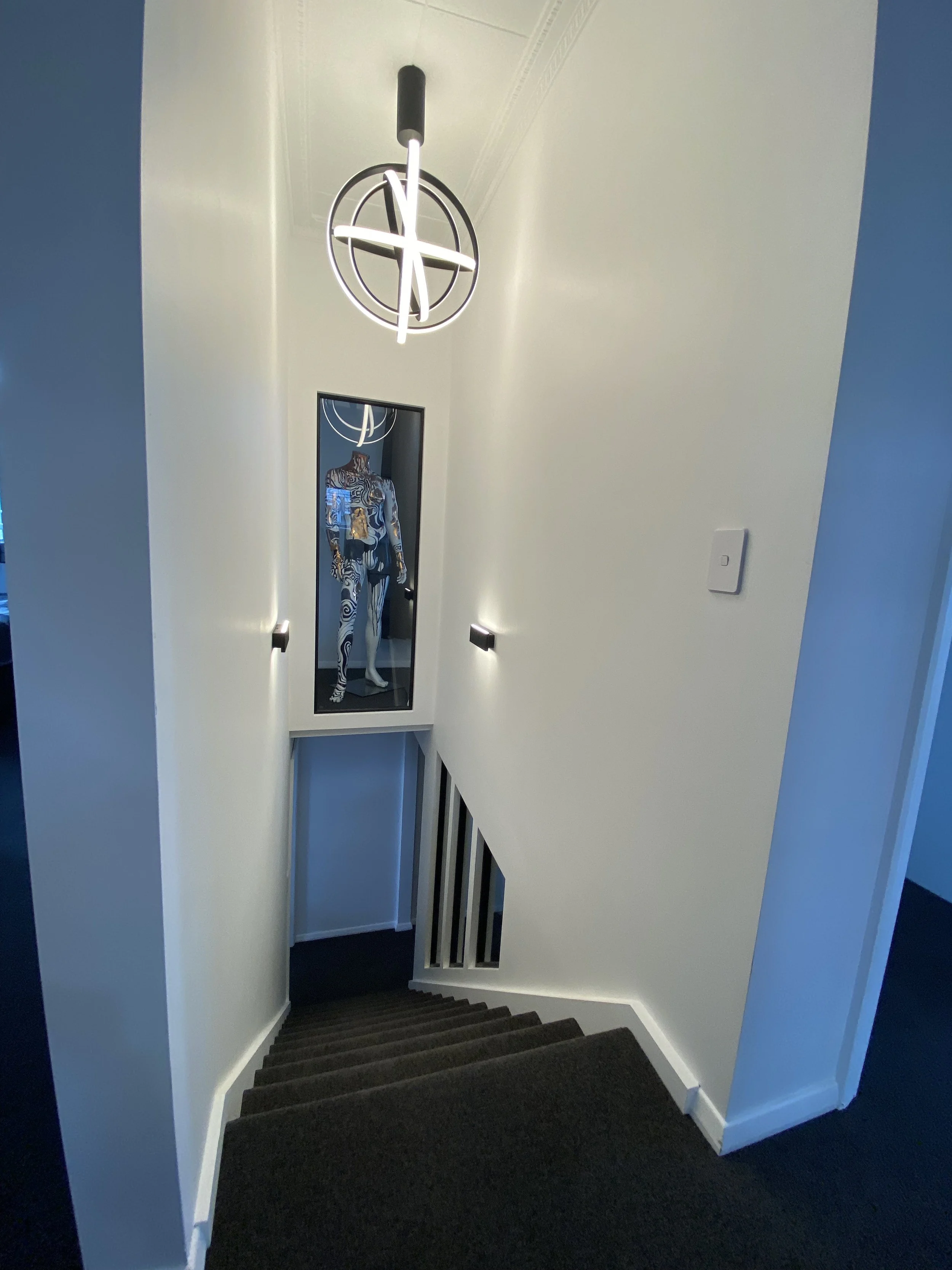 Interior view of a staircase leading down, with a modern geometric pendant light fixture hanging from the ceiling and a mannequin dressed in a blue and white patterned outfit visible at the bottom.