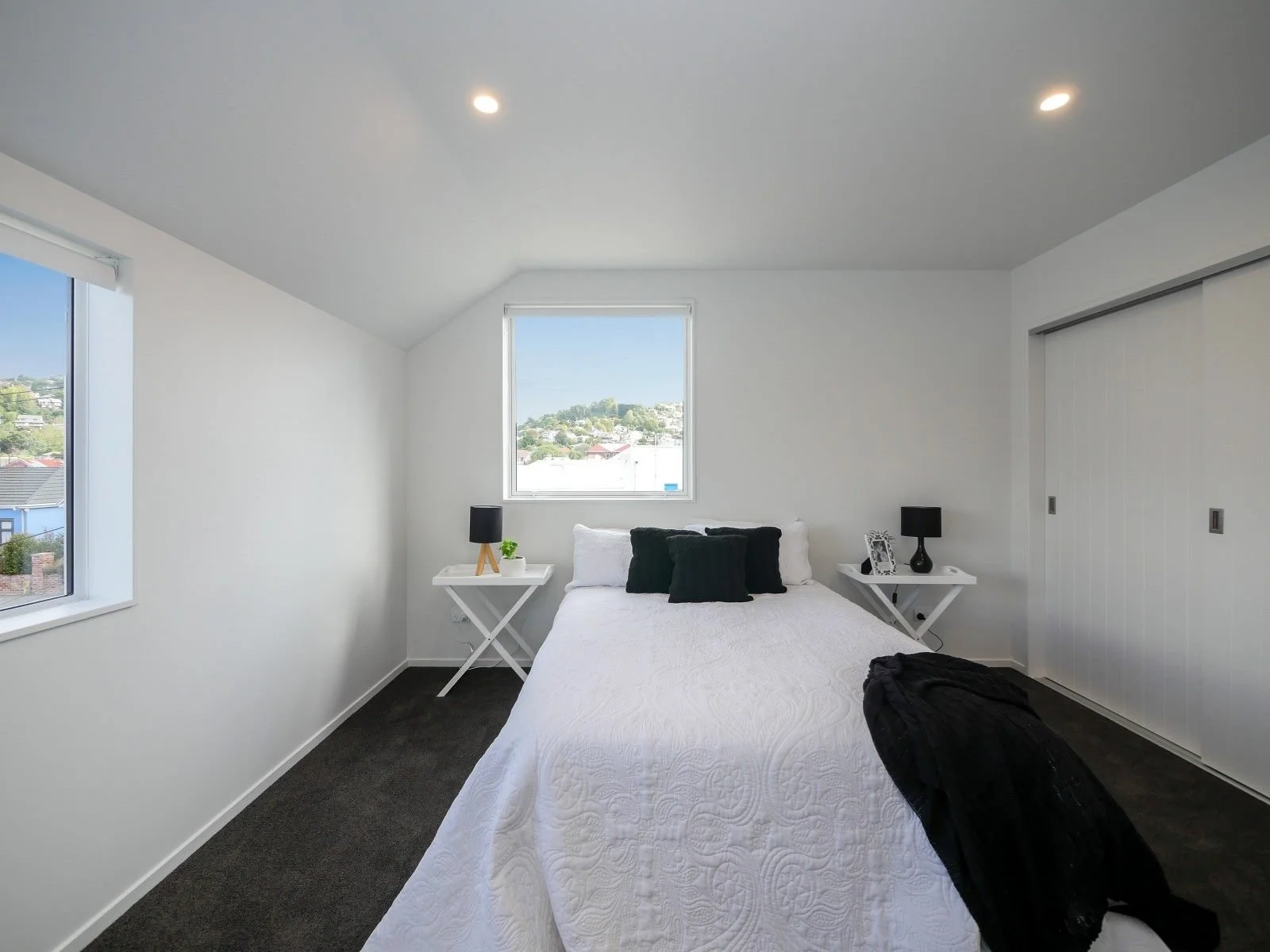 A bedroom with white walls, black carpet, a bed with white bedding and black pillows, two white nightstands with black lamps, a window showing a suburban neighborhood, and a closet with sliding white doors.