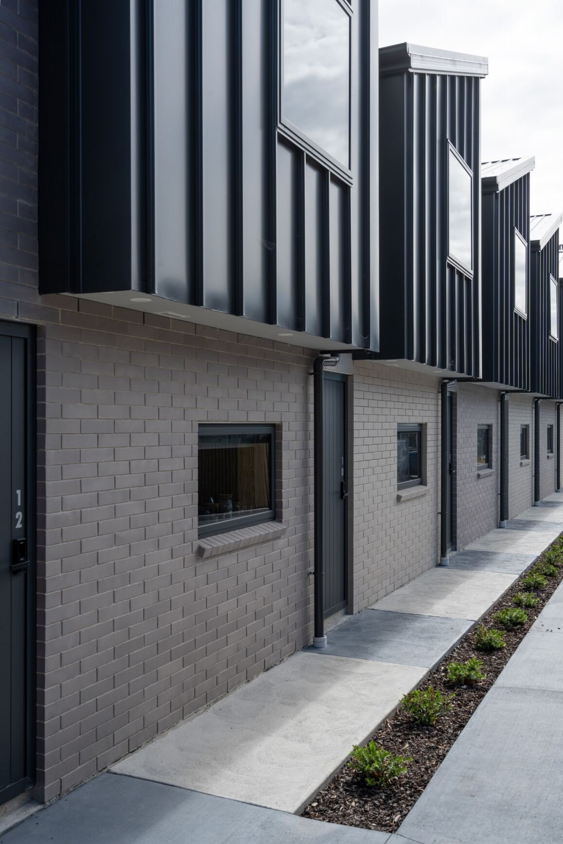 Modern row of townhouses with brick on the lower level and black metal siding on the upper level, featuring small windows and sidewalk gardens.