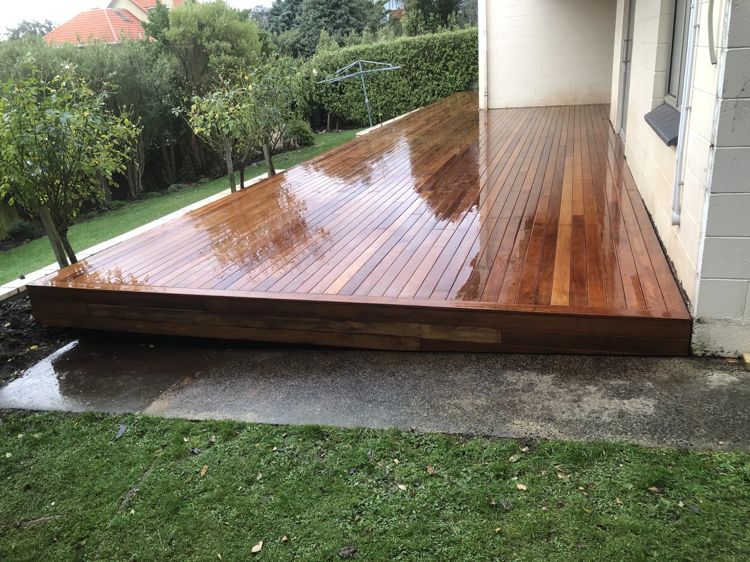Wooden deck with rain on it, adjacent to a grassy yard with trees and bushes, and a house with a brick wall and a window.