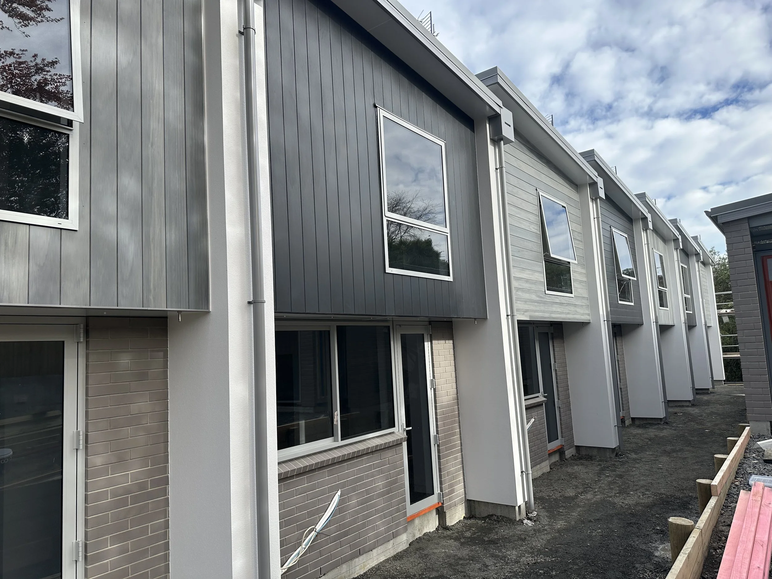 Row of modern townhouses under construction with large windows and exterior siding in shades of gray, with construction materials and dirt in the foreground.