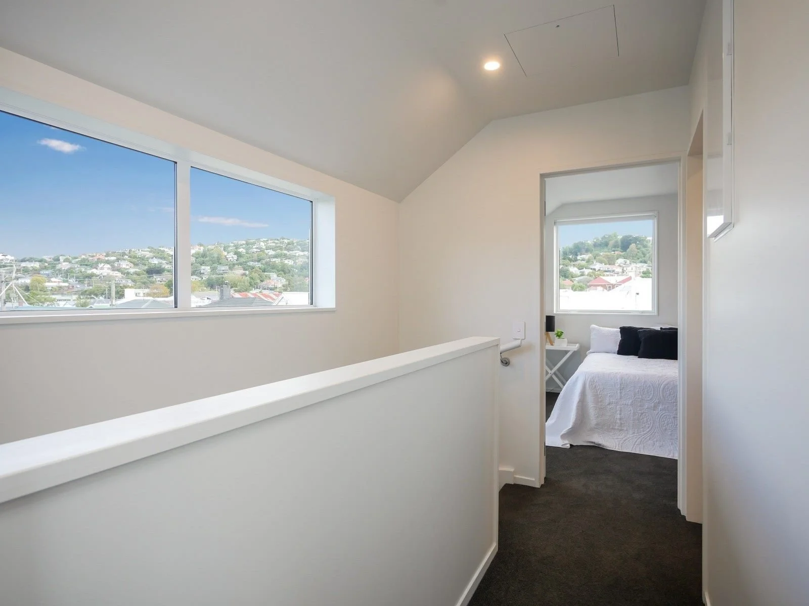 Interior view of a bright, minimalistic hallway with large windows showing a hilly landscape outside, leading to a cozy bedroom with a white bed and black pillows.