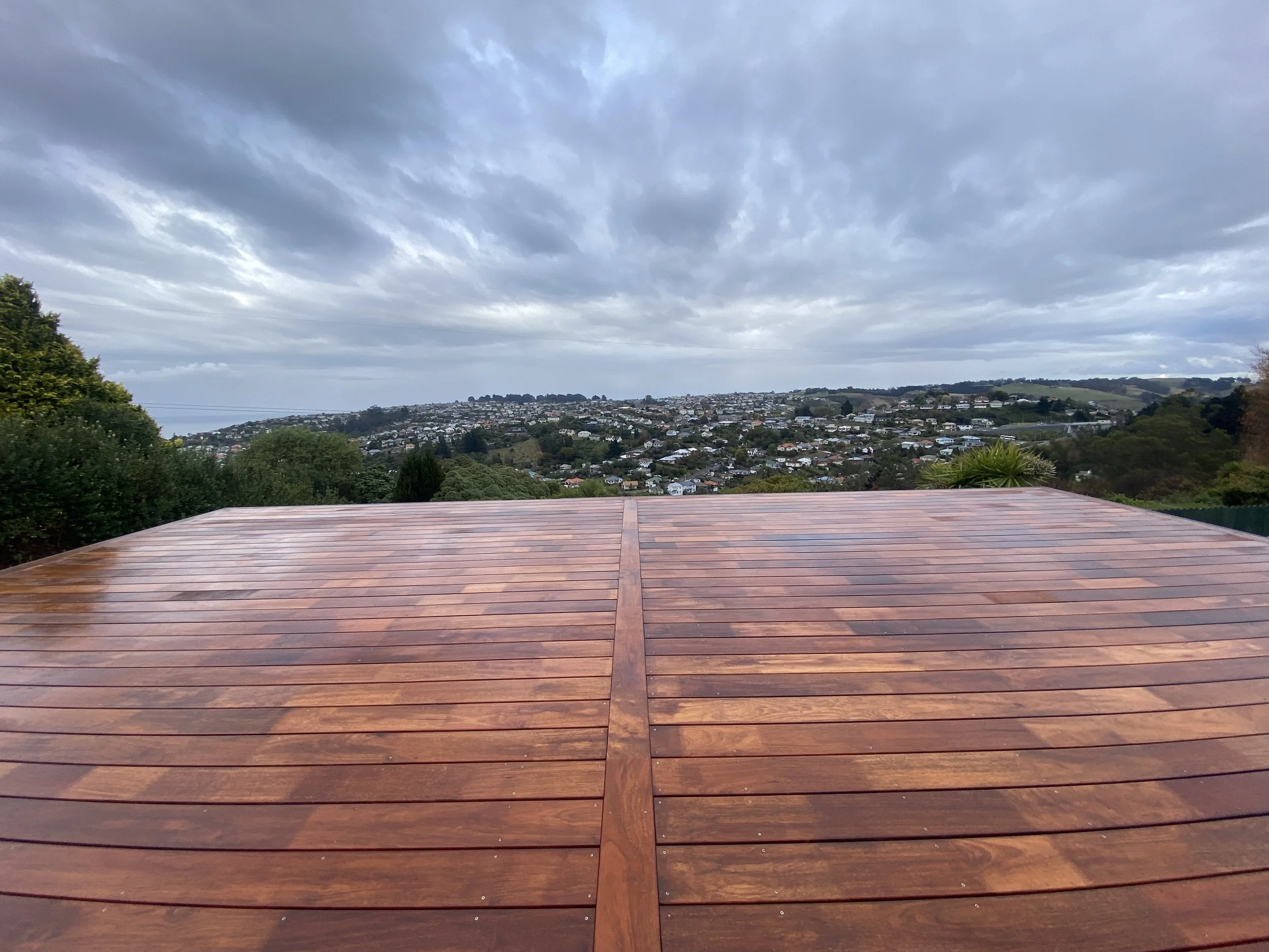 A view of a cloudy sky over a hillside neighborhood with houses and greenery, seen from a wooden deck in the foreground.