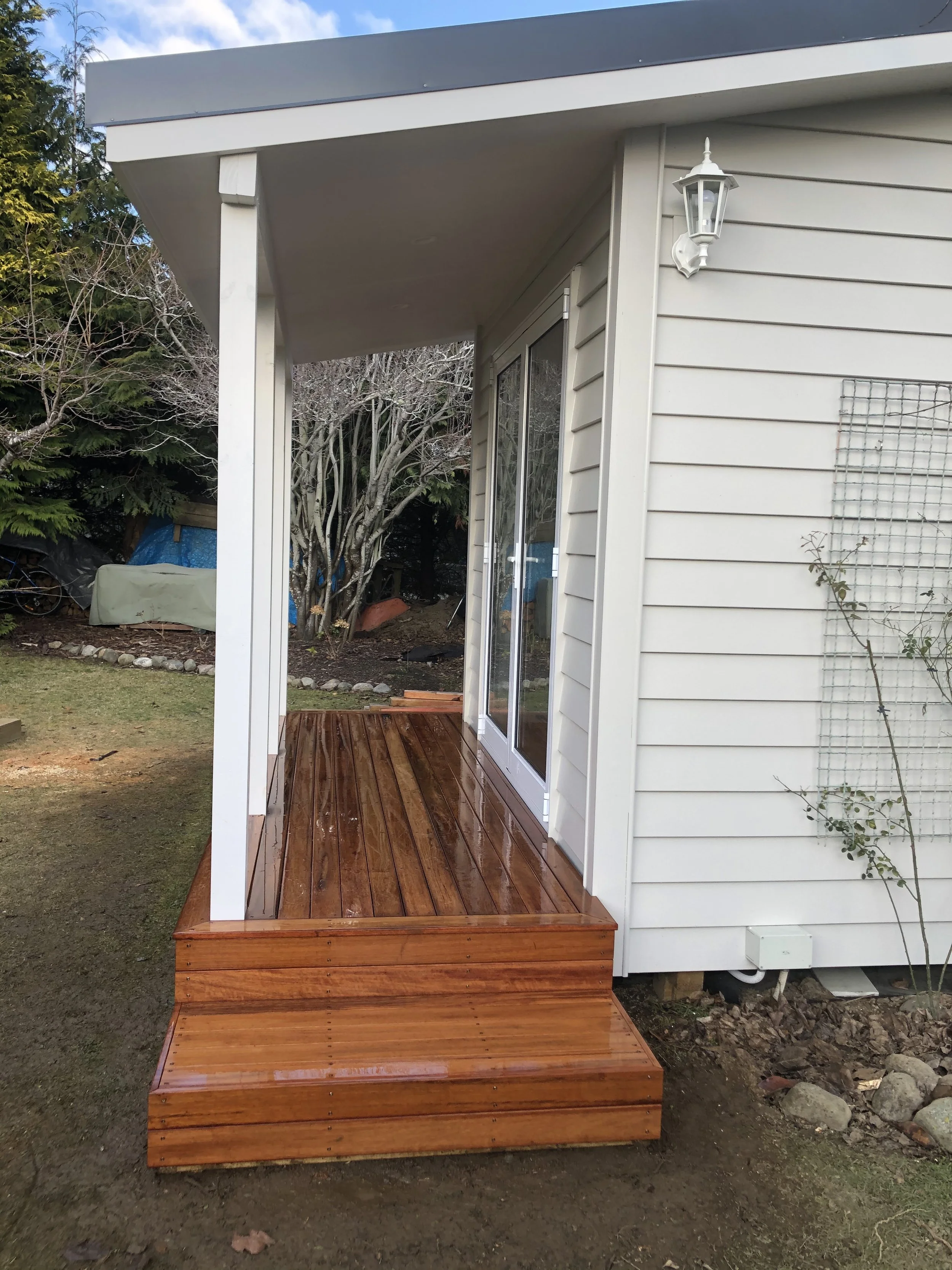 A small wooden deck attached to the back of a house with white exterior siding. The deck is next to a sliding glass door and has a roof overhang. There is a lantern-style outdoor light fixture mounted on the house wall. In the background, there are t