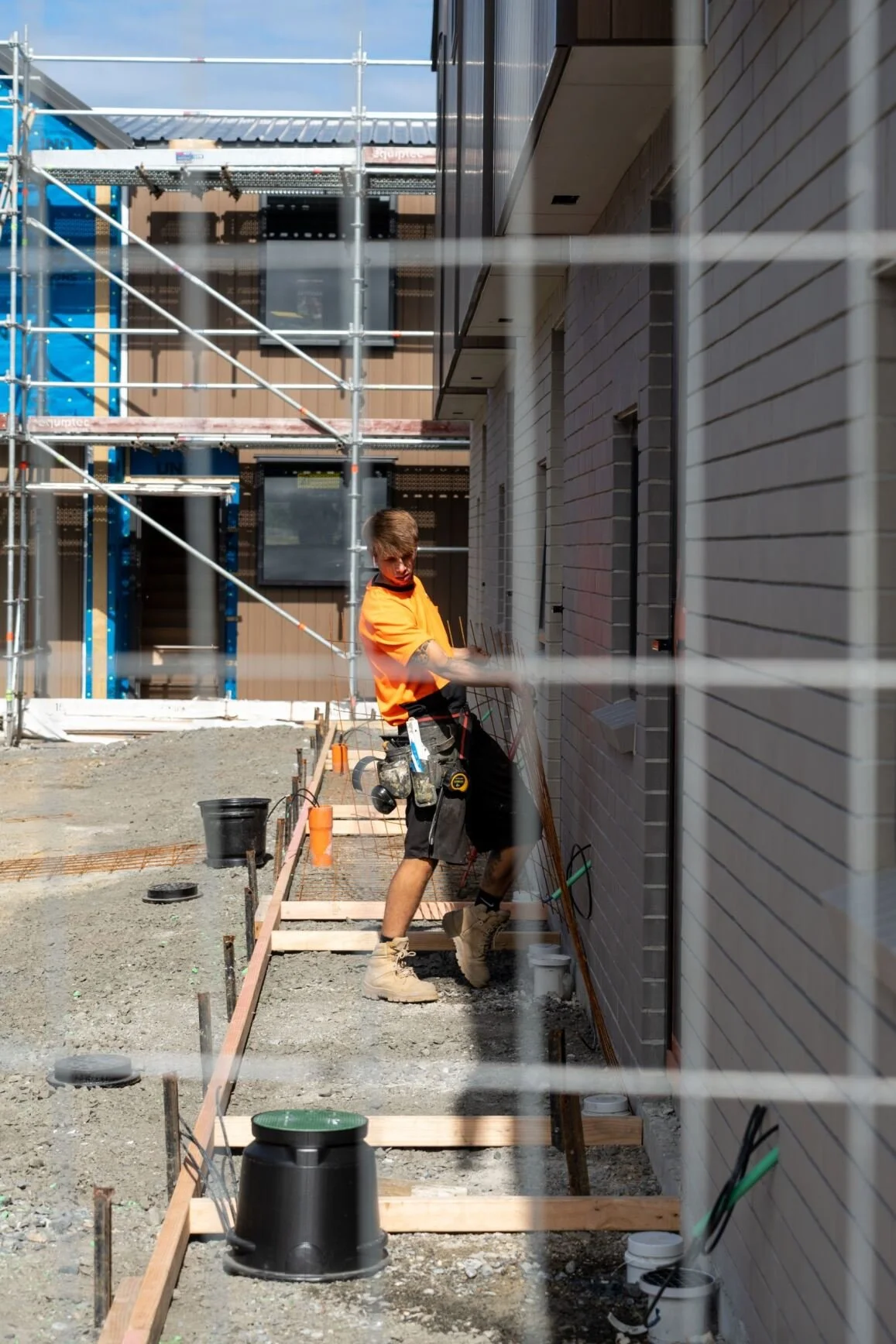 Construction worker in orange shirt and black shorts working on a building site, with scaffolding, pipes, and building materials around him.