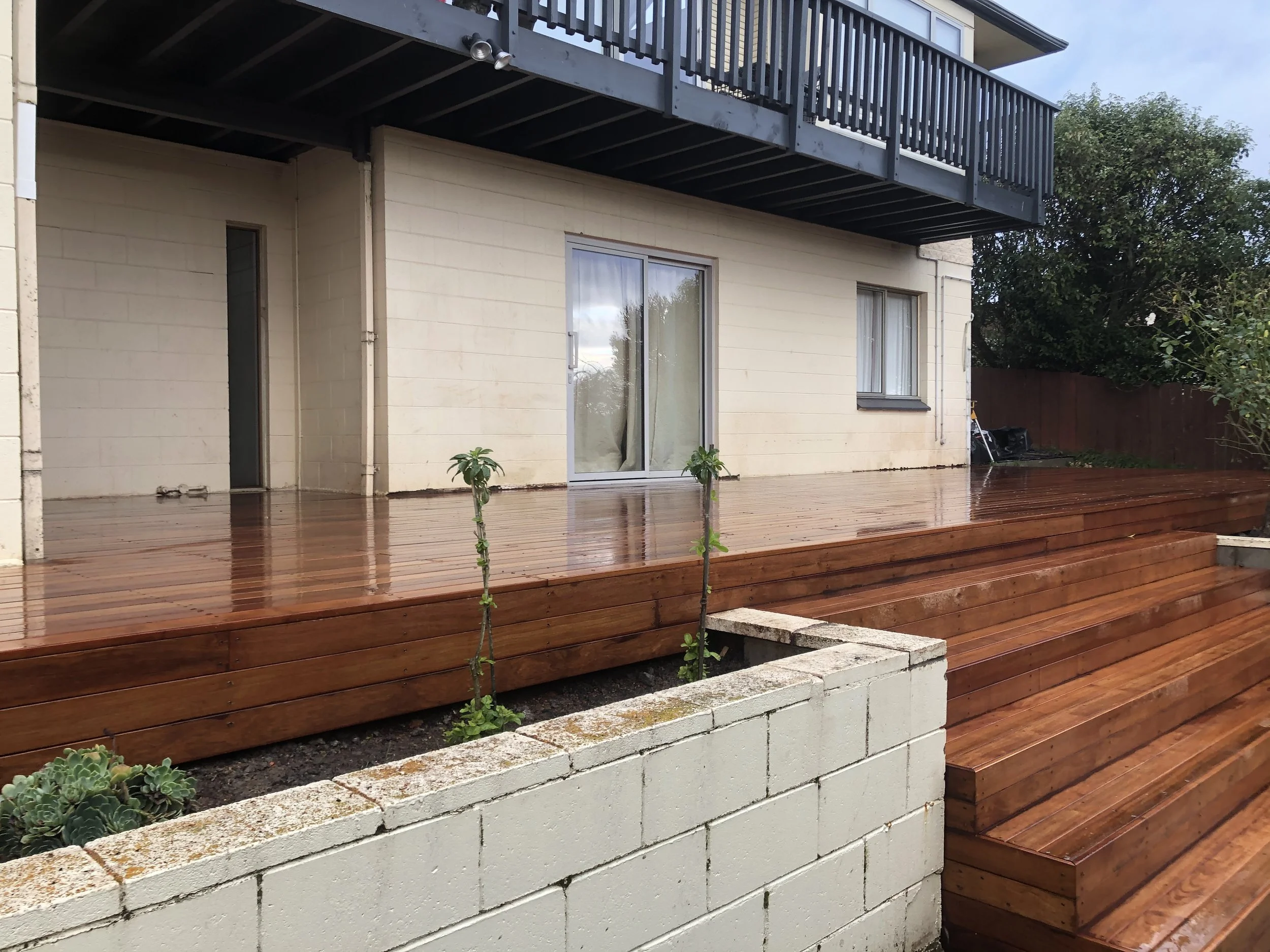 Backyard with wooden deck, small plants, white brick planter, sliding glass door, window, balcony with black railing, and trees in the background.