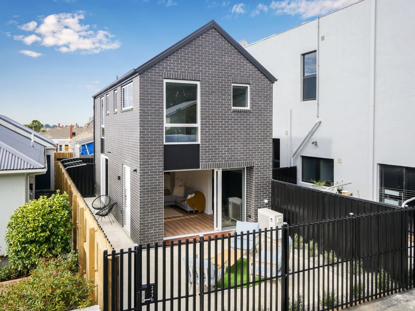 Modern multi-story residential building with dark gray brick exterior, large windows, and a backyard patio enclosed by a black fence. The patio contains outdoor seating and a small lawn area.